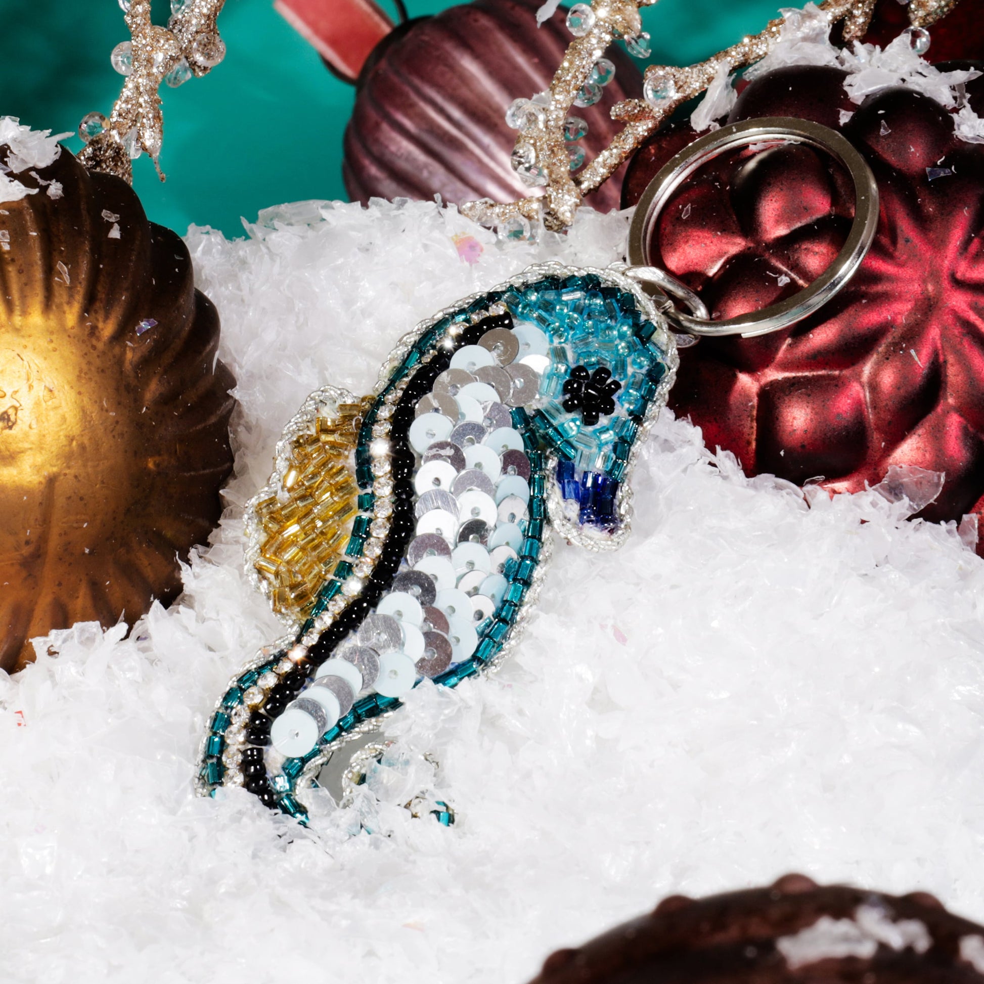 A beaded seahorse keyring decorated with turquoise, silver, and gold sequins resting on artificial snow among red and gold Christmas baubles and glittering gold branches against a teal background.