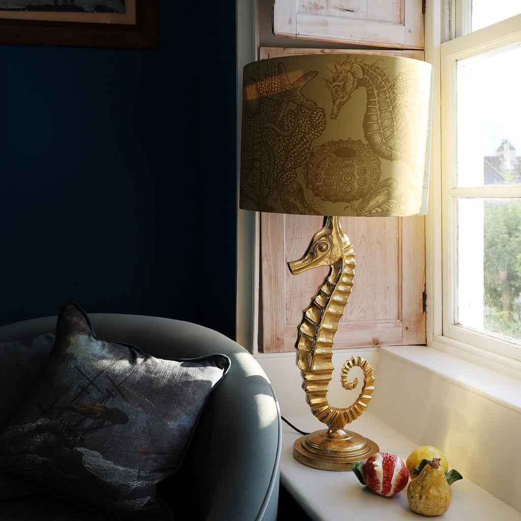 Gold seahorse lamp with decorative shade on a windowsill next to a cushioned chair.