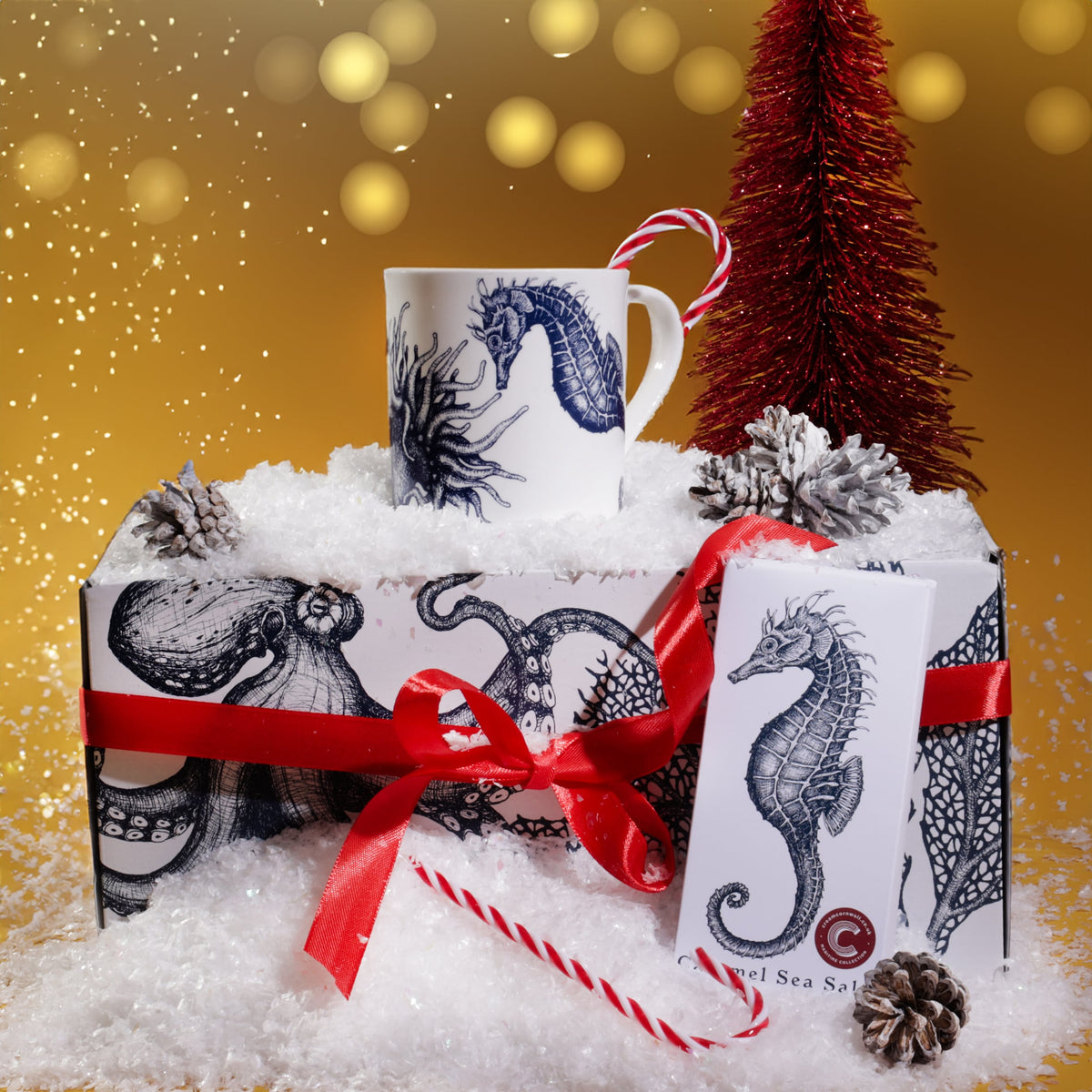 Seahorse mug and chocolate gift set displayed in artificial snow with red ribbon, candy cane, pinecones, and a red Christmas tree, set against a golden festive background with softly falling snow and glowing holiday lights.