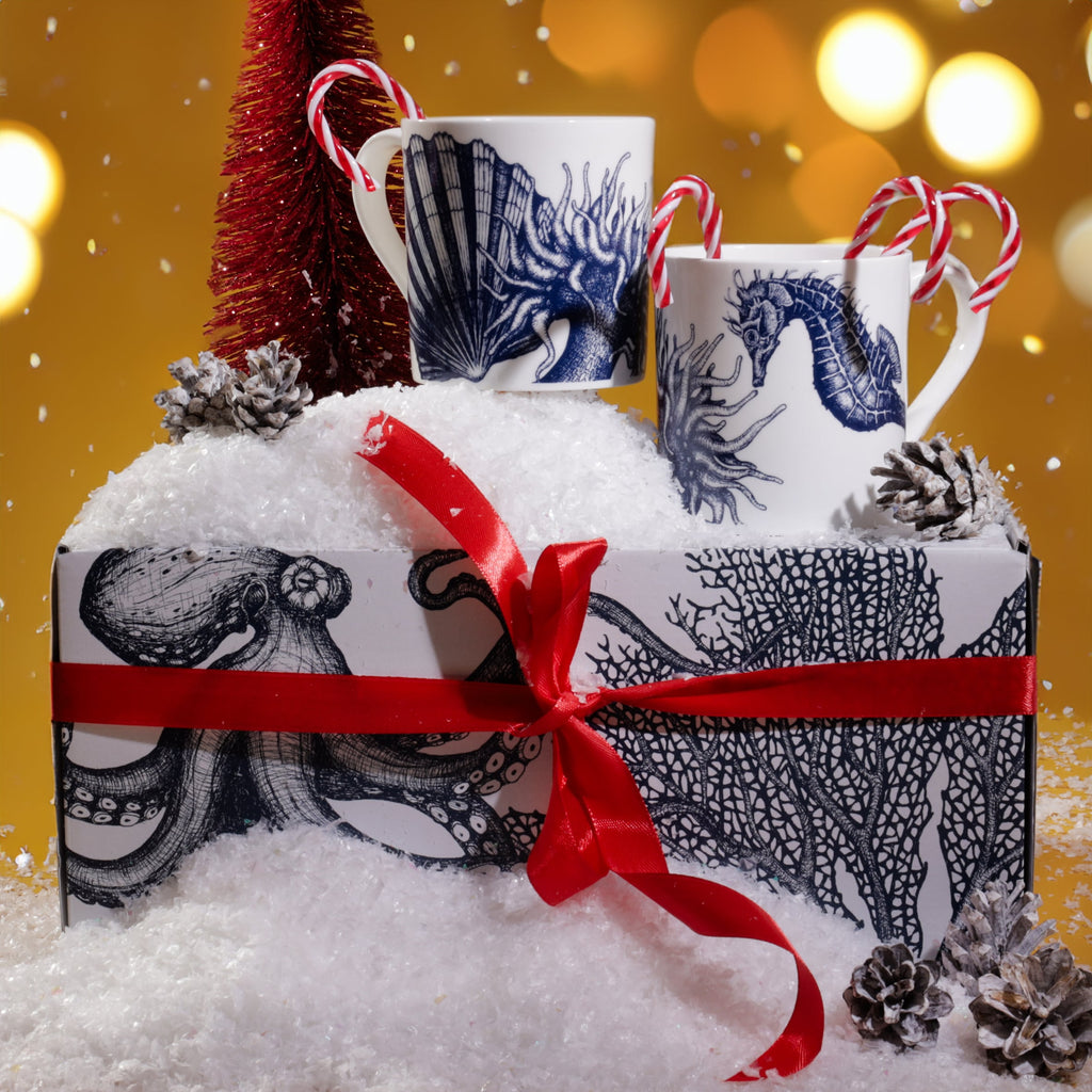 Seahorse mug gift set arranged in artificial snow with red ribbon, pinecones, candy canes, and a small red Christmas tree, against a warm golden background with twinkling festive lights.