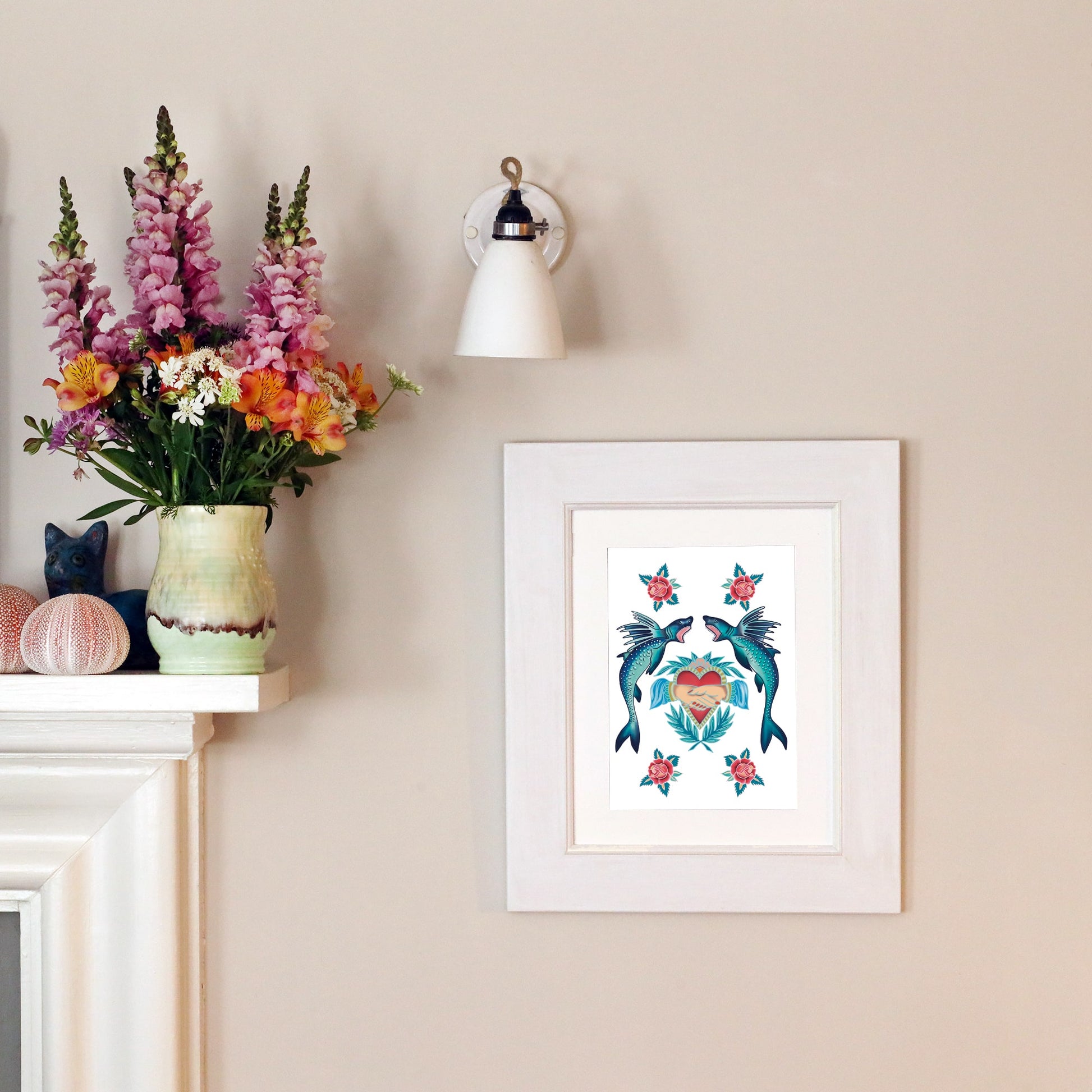 A framed tattoo-style print of two fish, a heart, and roses hangs on a pale wall beneath a white wall sconce. To the left, a mantel holds a vase of colorful flowers, decorative sea urchin shells, and a ceramic cat figurine.