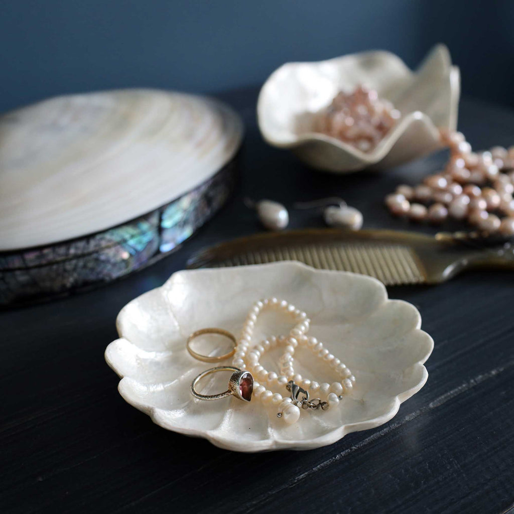 A scalloped ceramic jewelry organizer tray in the foreground holding pearl strands, rings, and a small charm, with additional pearl accessories and a wooden comb on a dark tabletop. A larger shell-shaped dish and abalone-embossed box are blurred in the background.