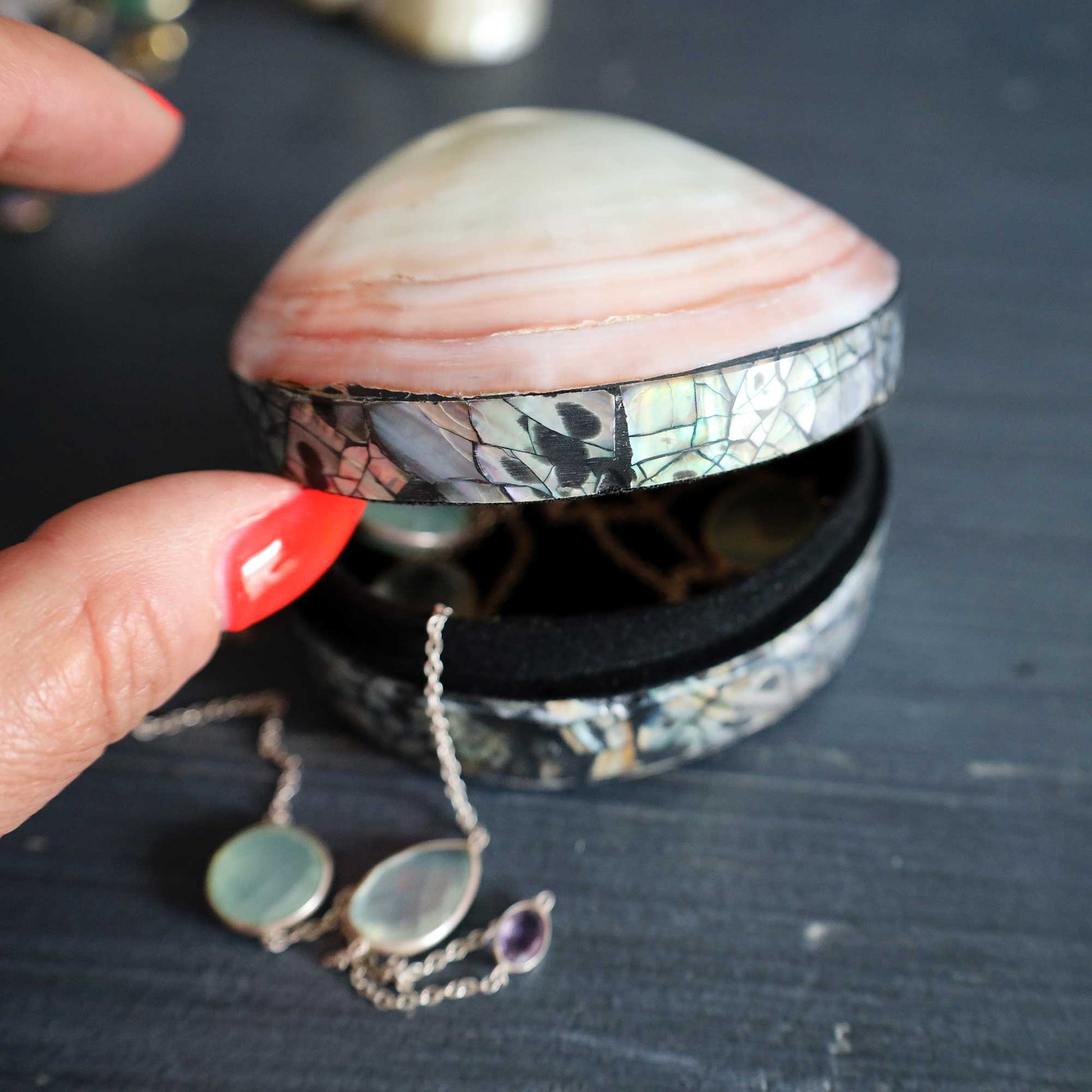 A handmade clam-shell jewelry box with iridescent mother-of-pearl inlay, shown open with a necklace resting inside, held by a hand with red nail polish on a dark wooden surface.