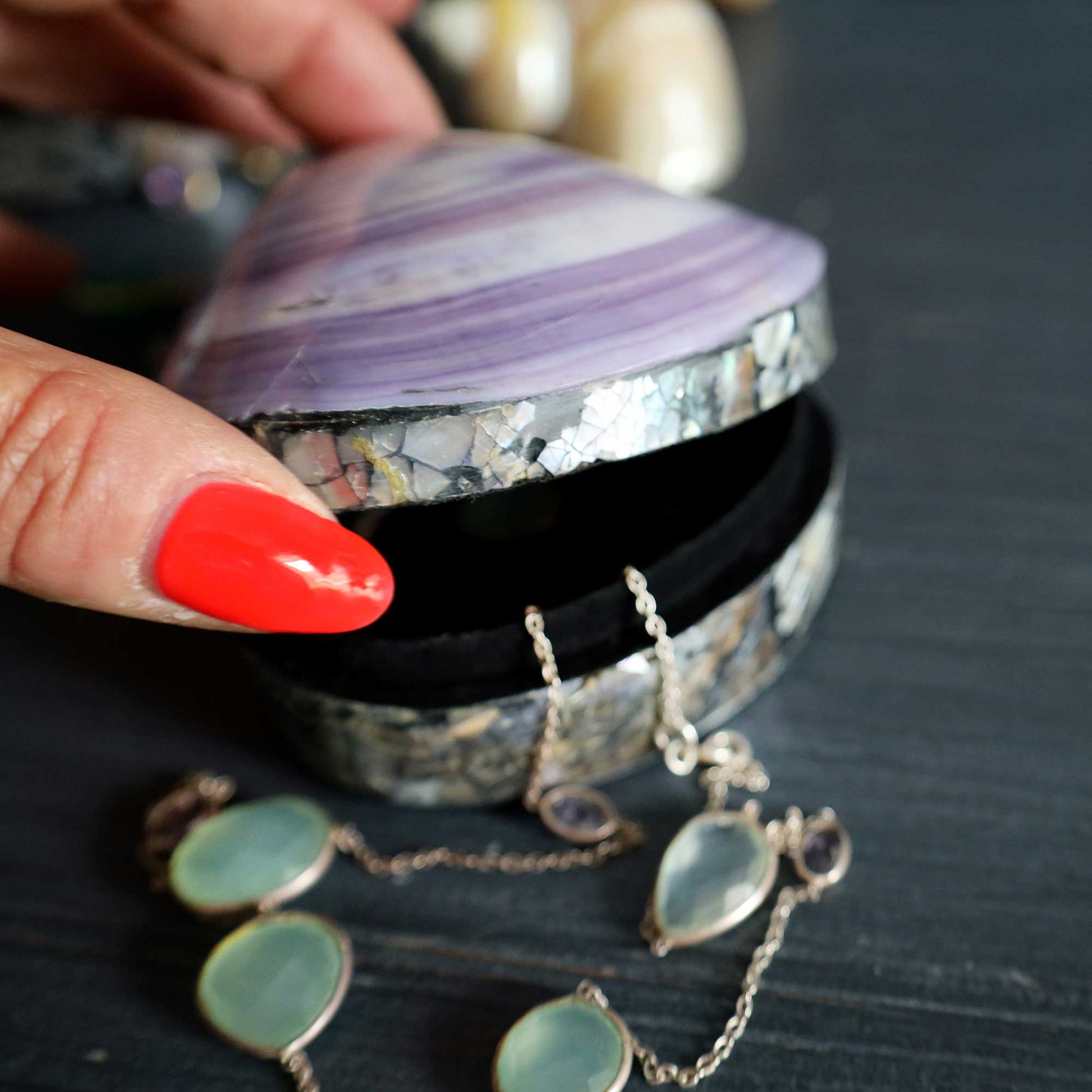 A close-up of a hand with bright red nail polish opening a shell-shaped jewelry box with a purple and white striped interior. Silver-toned jewelry with pale green and purple stones is draped out of the box on a dark tabletop.