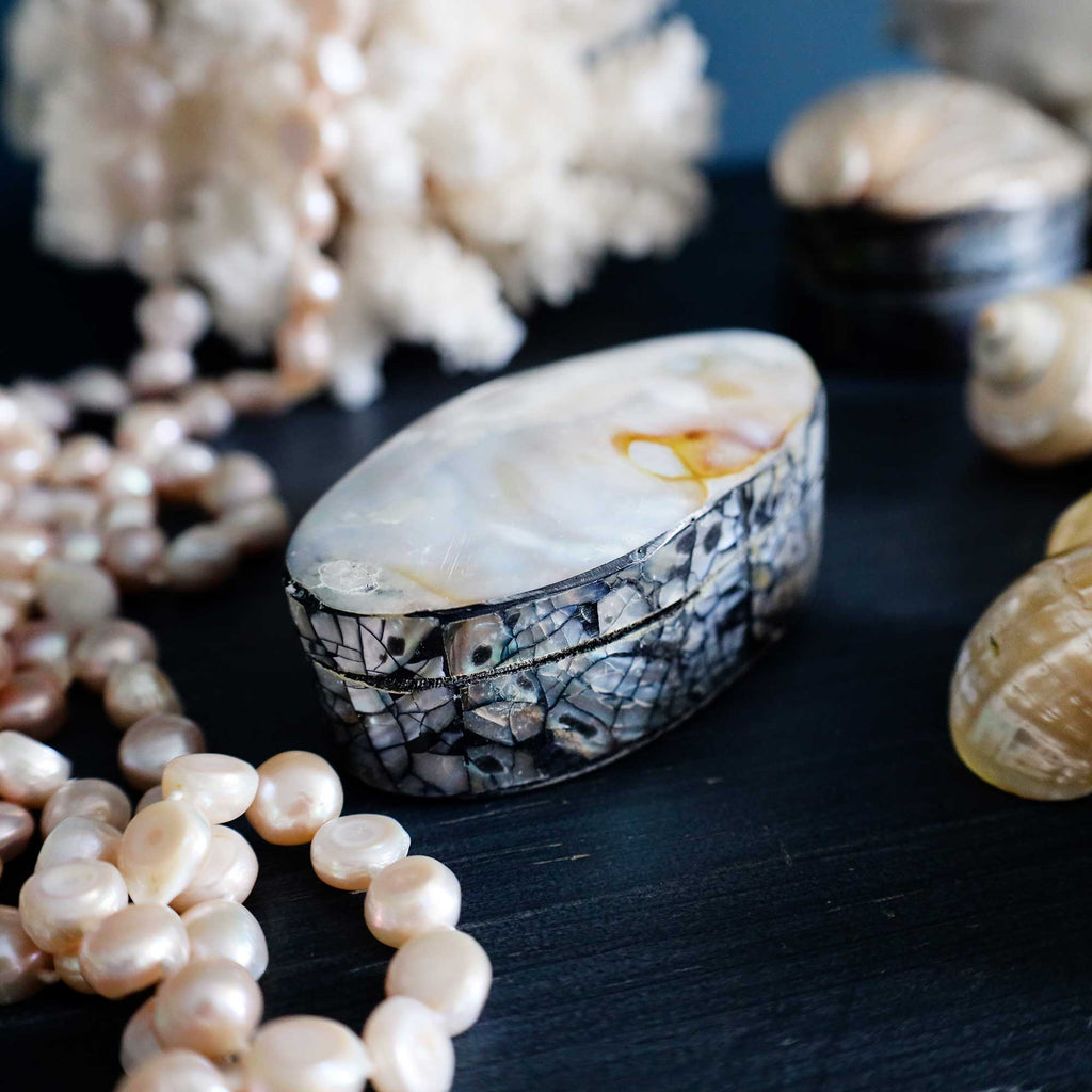 An oval shell-look jewelry box with a mosaic shell patterned exterior and a pearlescent cream interior, styled on a dark surface with pearls, shell fragments, and seashells around it.