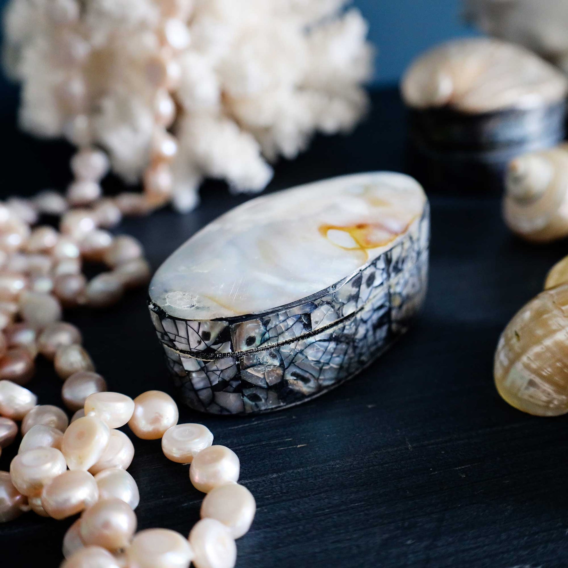 An oval shell-look jewelry box with a mosaic shell patterned exterior and a pearlescent cream interior, styled on a dark surface with pearls, shell fragments, and seashells around it.