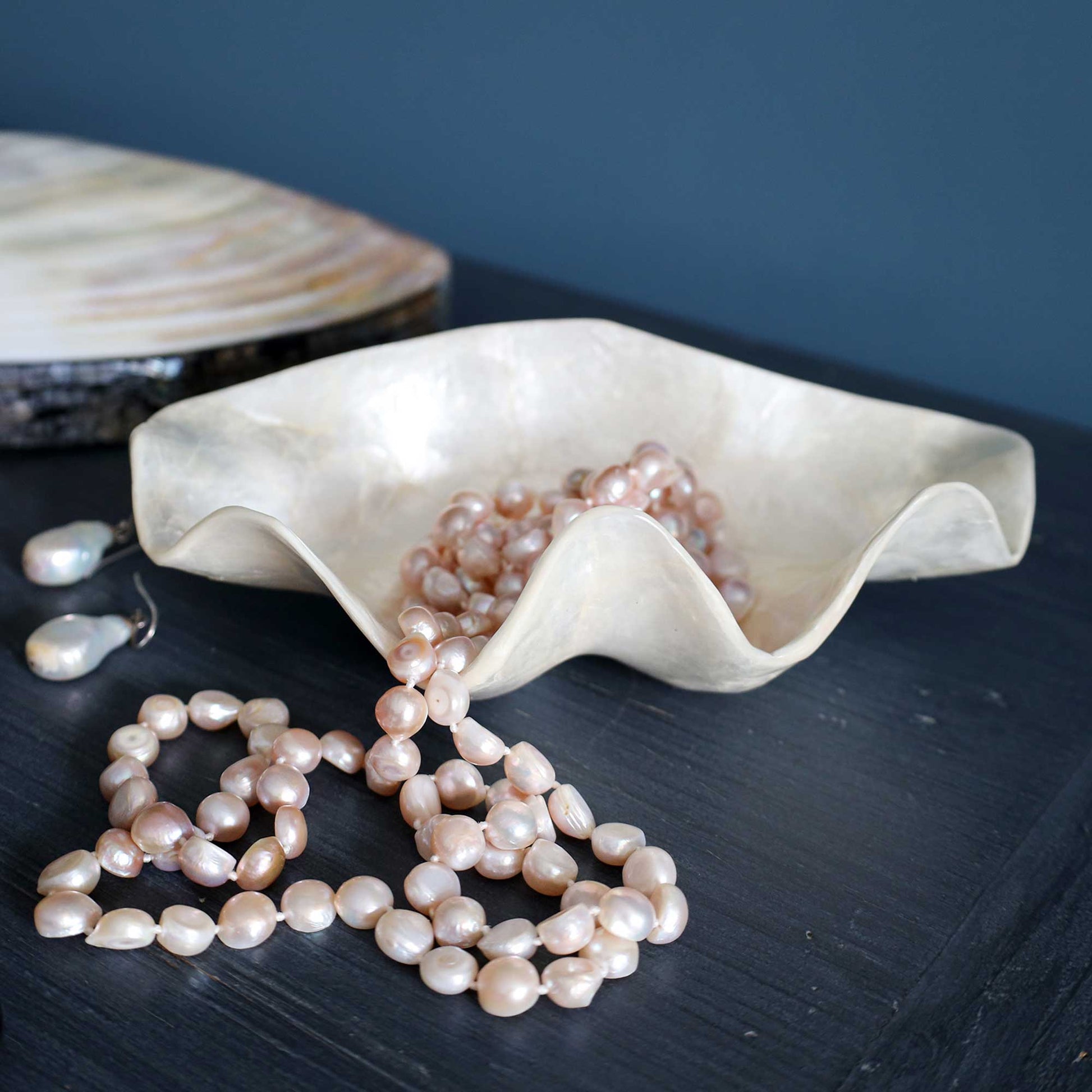 A shell-shaped cream-colored bowl filled with and draped over a strand of baroque pearls, with matching pearl earrings resting on a dark wooden surface against a dark blue background.
