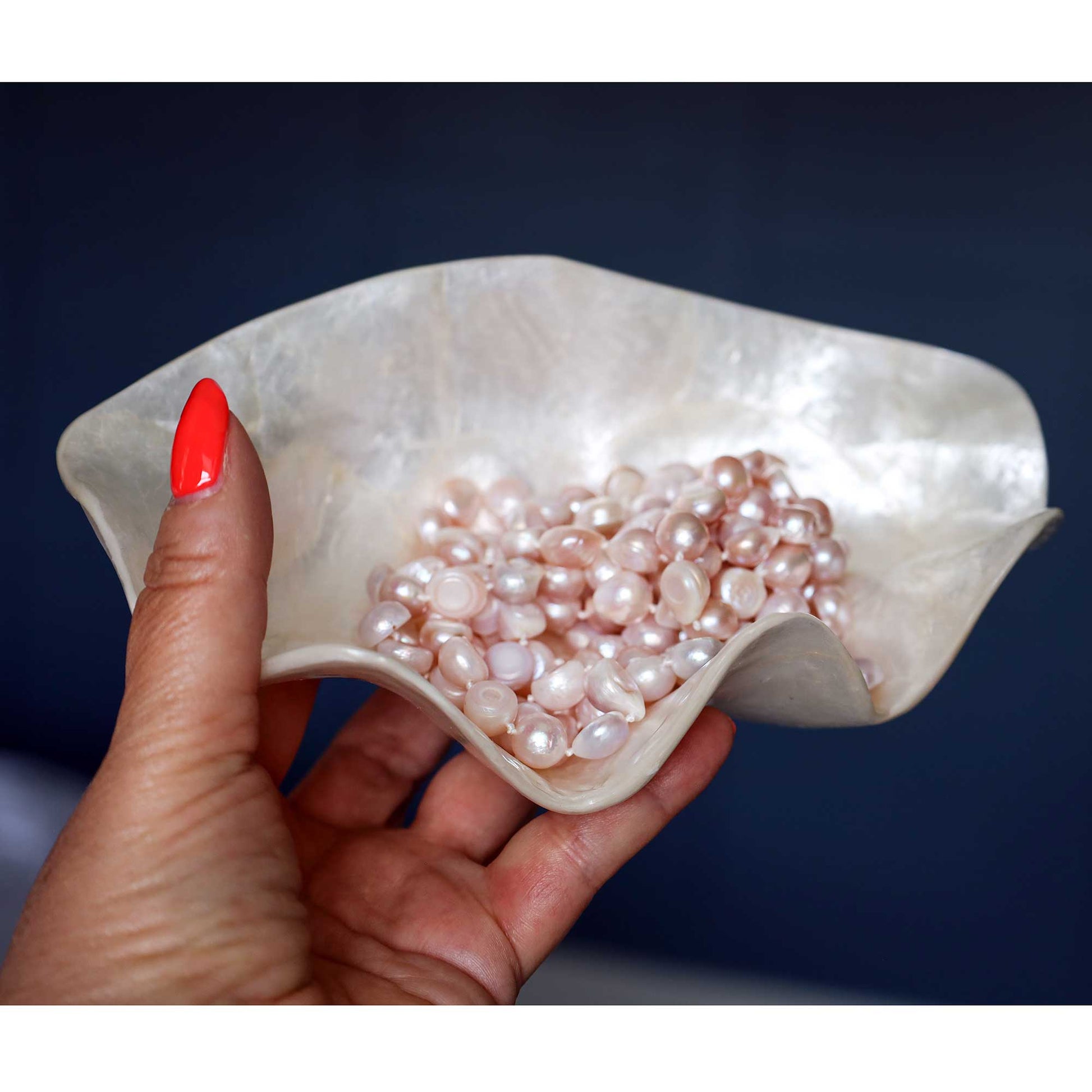 A hand with bright red nail polish holds a wavy, shell-like dish filled with pale pink baroque pearls against a dark navy blue background.