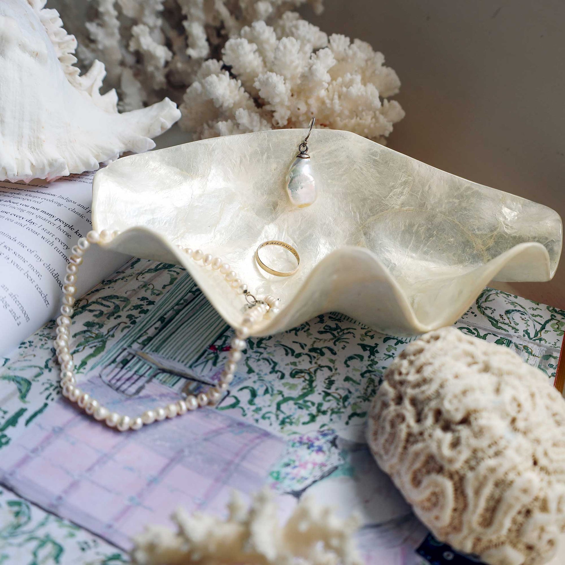 A seashell-shaped jewelry organizer holding pearl necklaces, rings, and a pendant, styled with coral, a knit blanket, and a book.