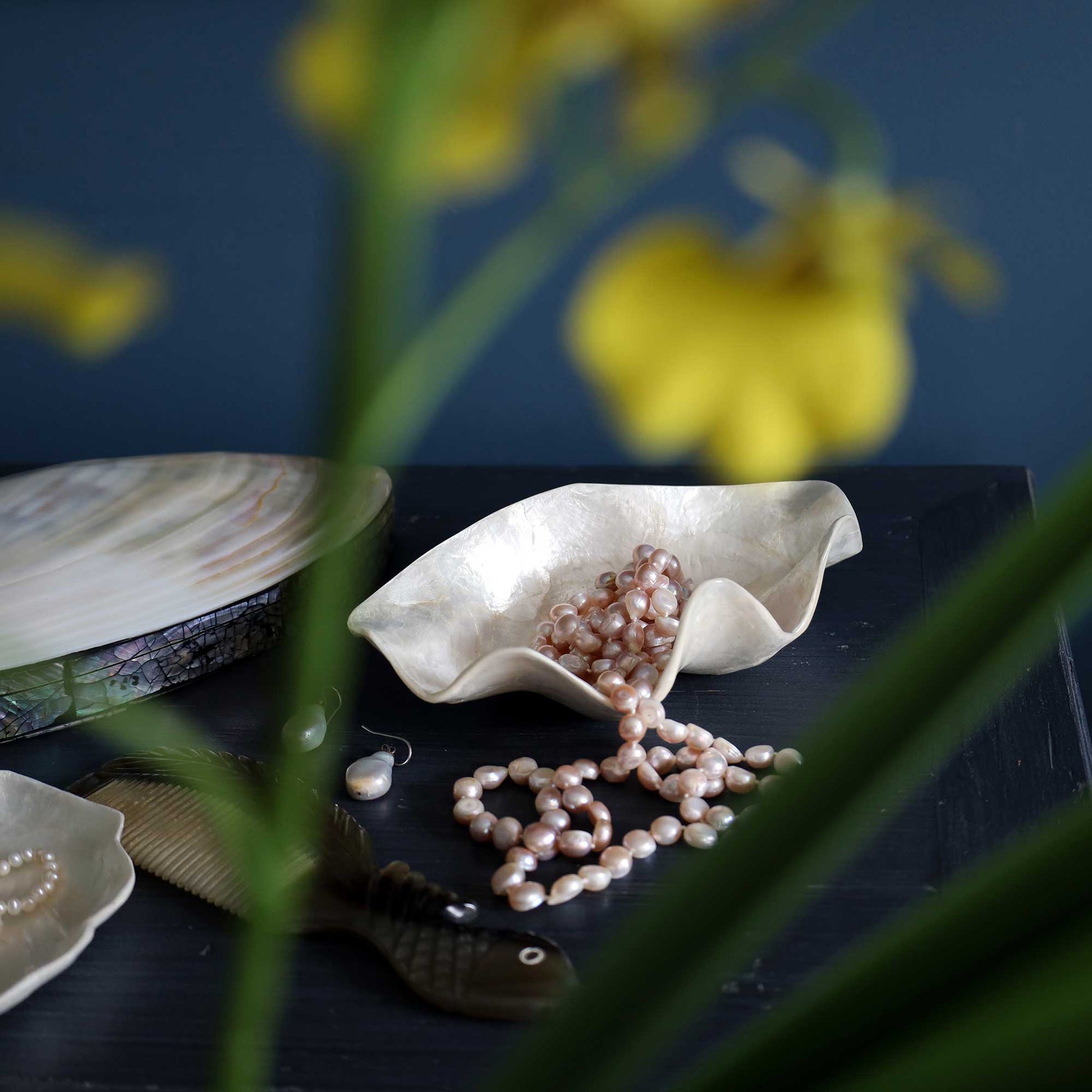 A styled still life featuring a shell-shaped dish filled with and spilling pale pink freshwater pearls, alongside a fish-shaped dish, a shell dish with abalone shell detailing, and a comb, all arranged on a dark tabletop with a dark blue background and blurred yellow flowers in the foreground.
