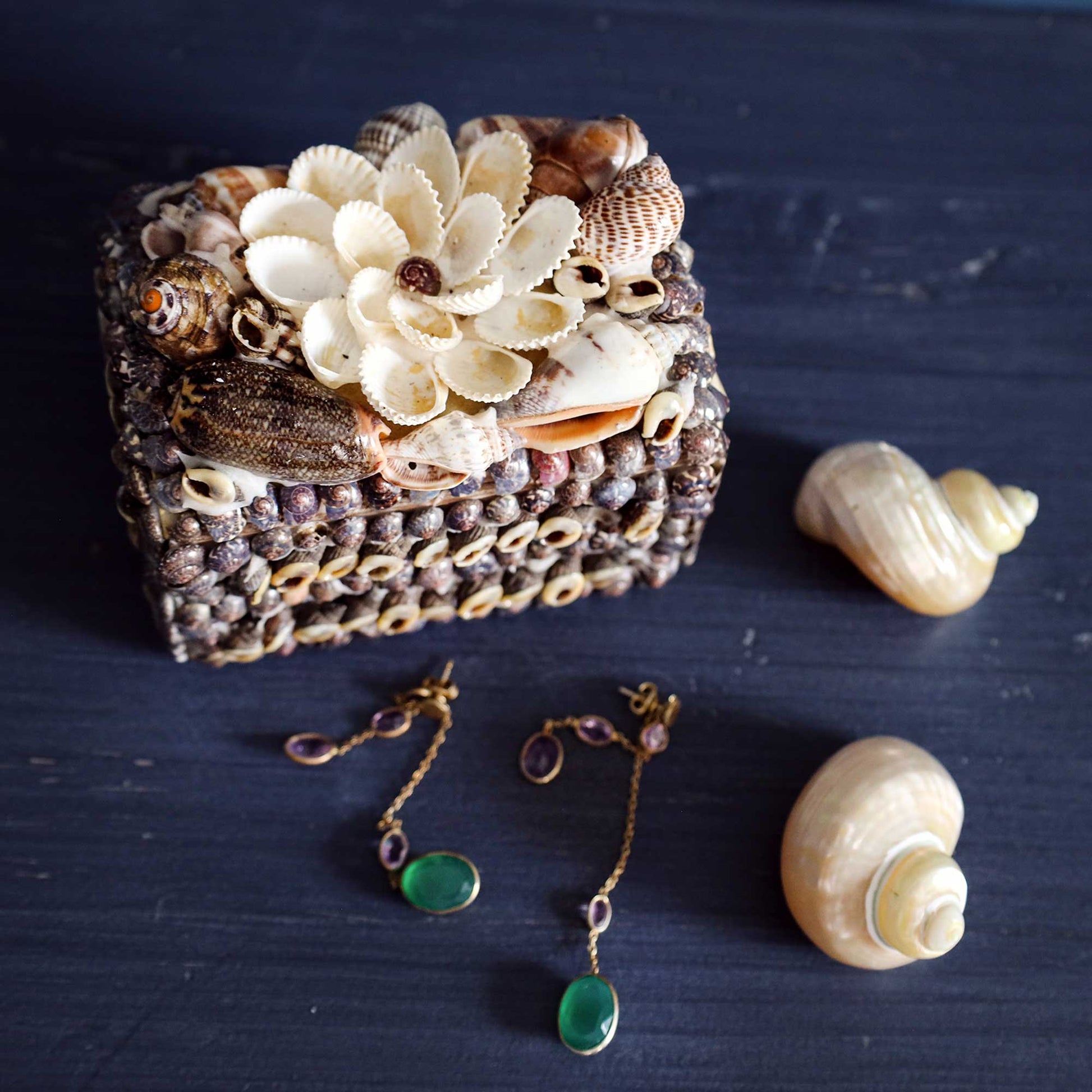 A decorative jewelry box made from seashells and small shells, styled on a dark blue wooden surface with matching seashell ornaments and dangling green bead earrings nearby.