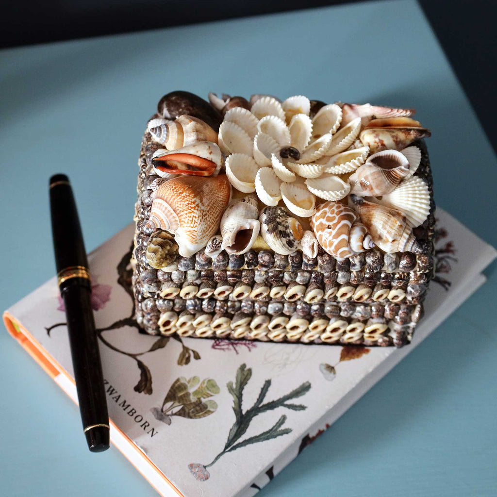 A rectangular decorative box covered with assorted seashells and shell flowers, placed on top of a floral notebook with a black pen beside it, on a light blue surface.