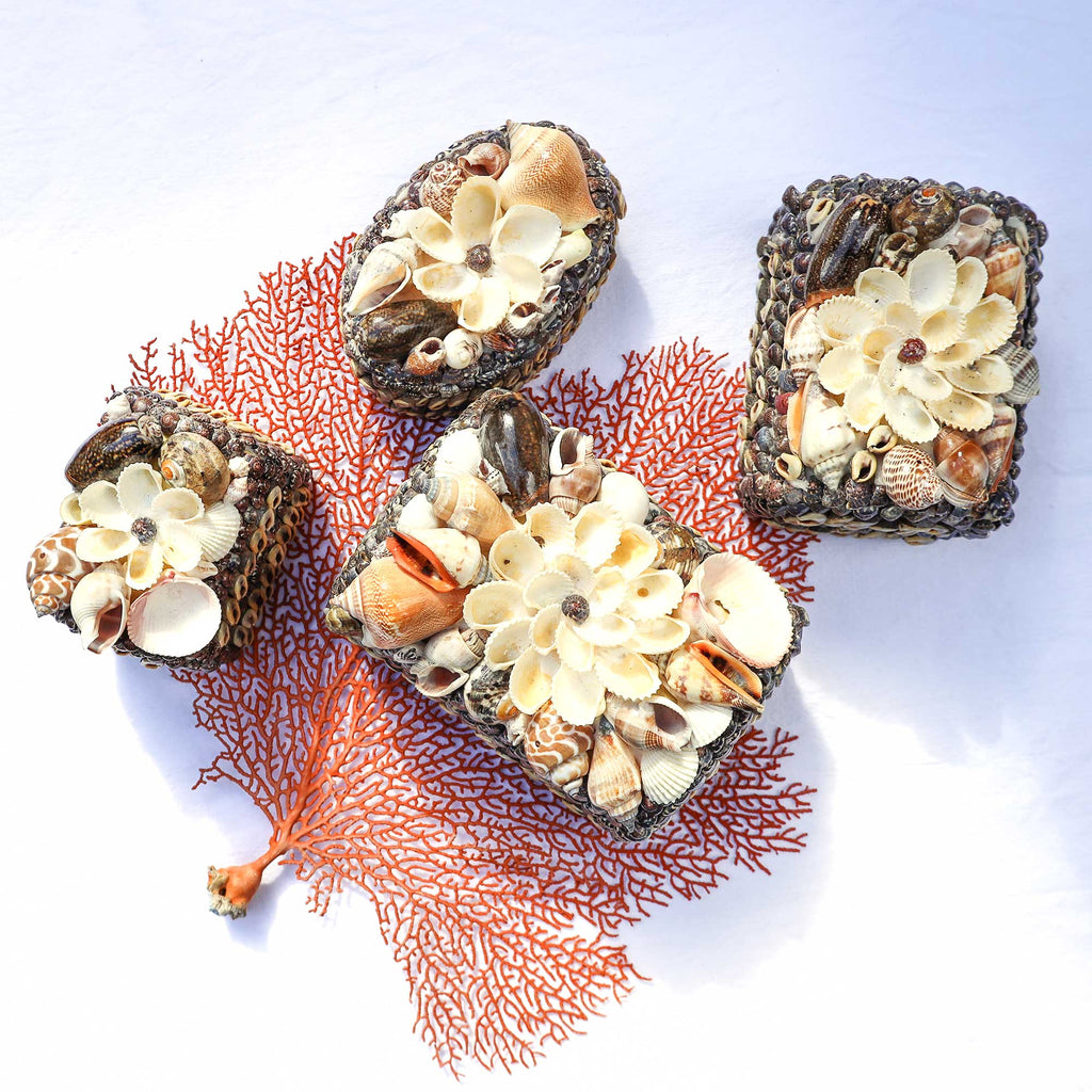 Three decorative rectangular and oval plaques made from dark woven material, embellished with cream and beige seashells and flower-shaped shell motifs, arranged on a white background with a coral branch prop.