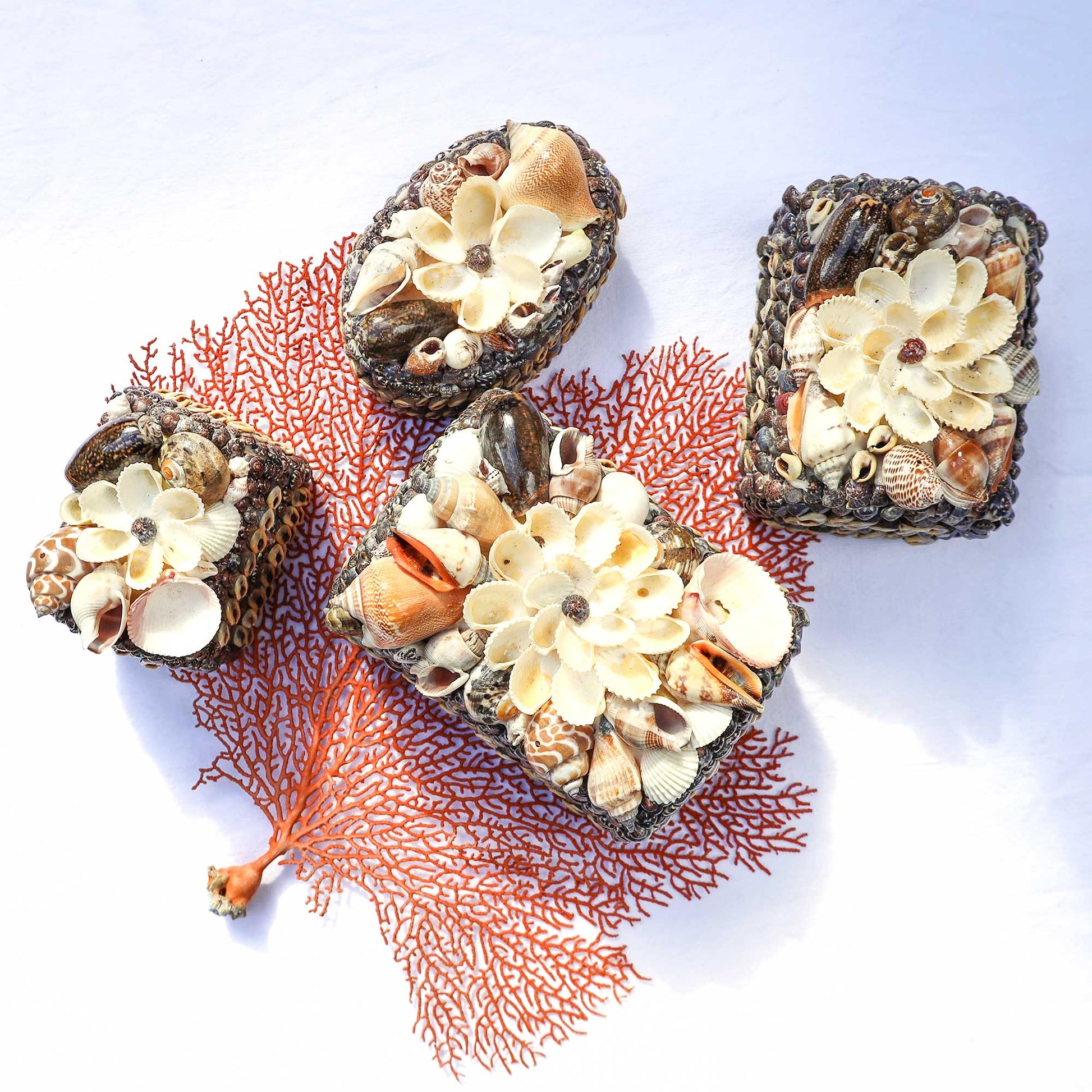 Three decorative rectangular and oval plaques made from dark woven material, embellished with cream and beige seashells and flower-shaped shell motifs, arranged on a white background with a coral branch prop.