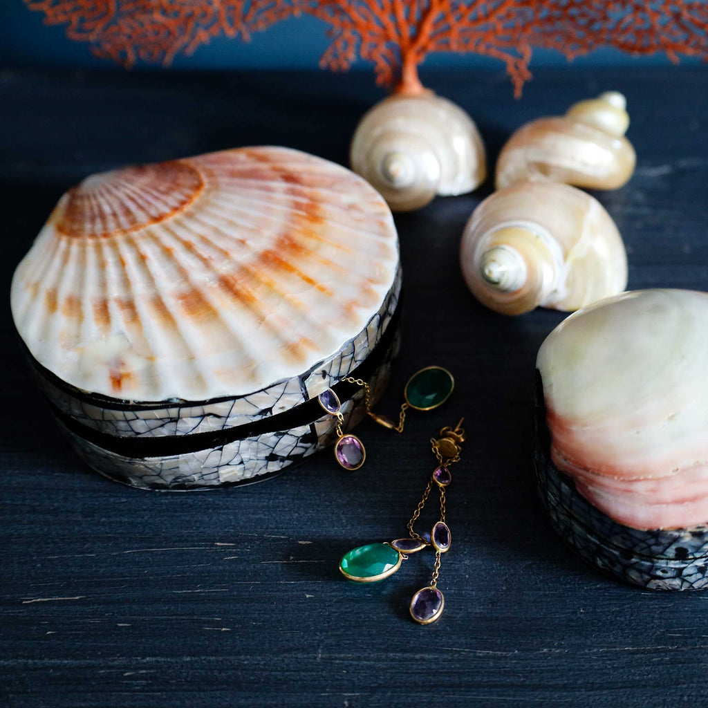 Gold-tone dangle earrings with green teardrop-shaped stones and purple rectangular stones, displayed on a dark wooden surface next to seashells.