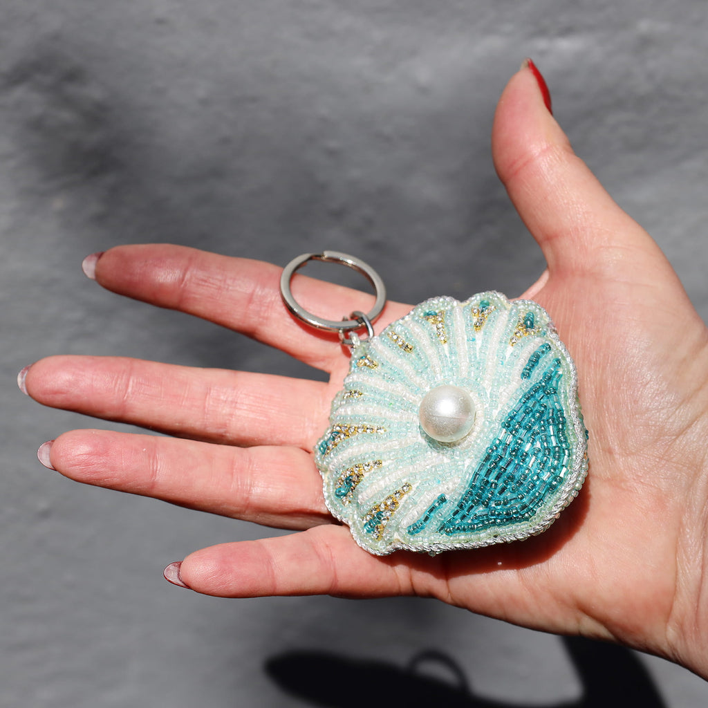 Hand holding a decorative keychain shaped like a shell with a pearl on a gray background