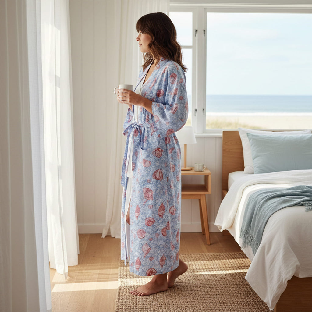 Woman in a floral robe holding a mug in a bedroom with a bed and window in the background.