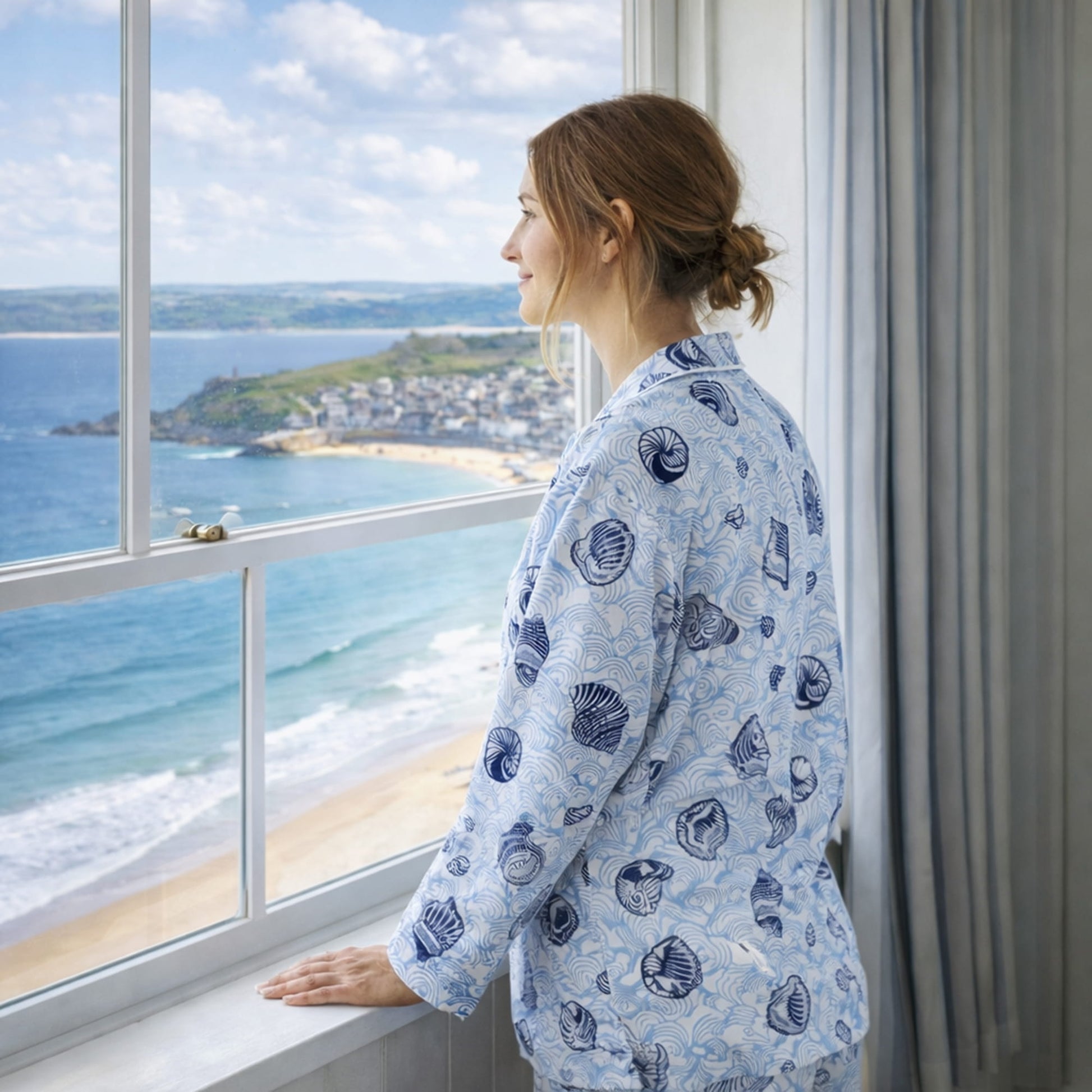 Woman in a blue patterned robe looking out of a window with a scenic view of the ocean and beach.