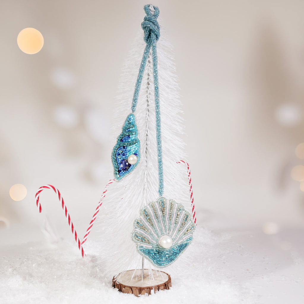 Two beaded shell bag charms in turquoise and blue tones with pearl details hanging from a small white Christmas tree, surrounded by artificial snow and candy canes with soft festive lights in the background.