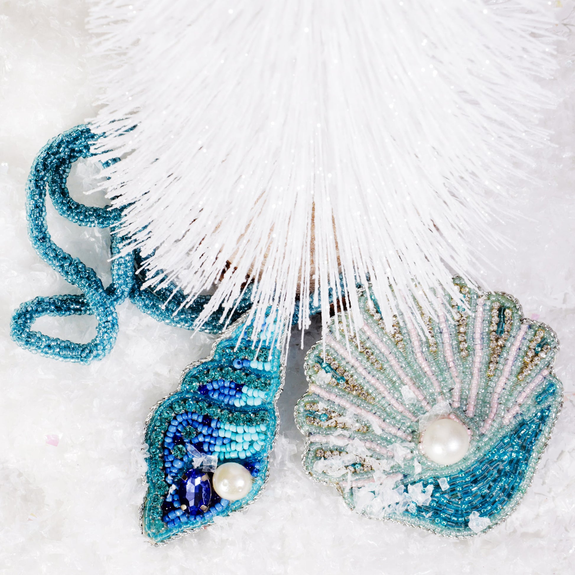 Beaded shell bag charms in shades of turquoise, blue, and gold with pearl accents displayed beneath a white Christmas tree, surrounded by artificial snow.