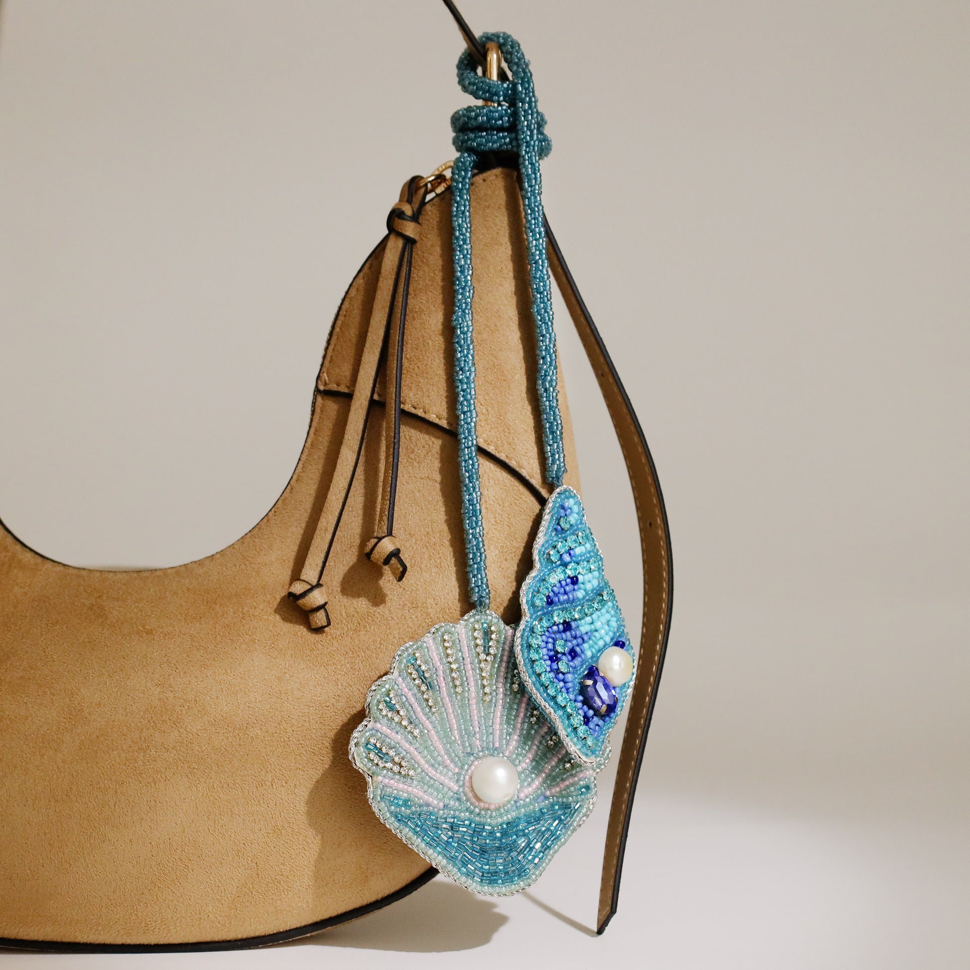 Two beaded shell bag charms in turquoise and blue tones with pearl details hanging from the handle of a tan suede handbag.
