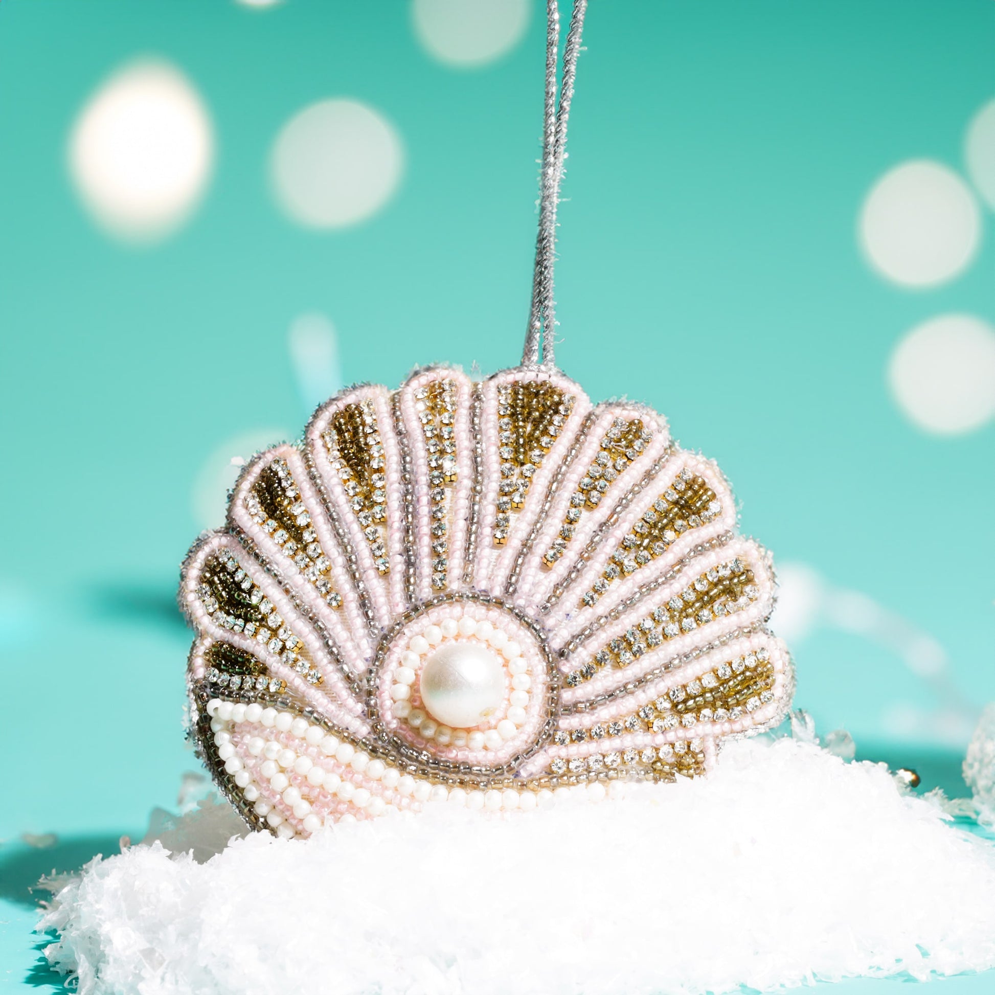 A beaded seashell decoration with gold, pink, and pearl details featuring a central faux pearl, resting on white snow against a turquoise background with soft glowing lights.