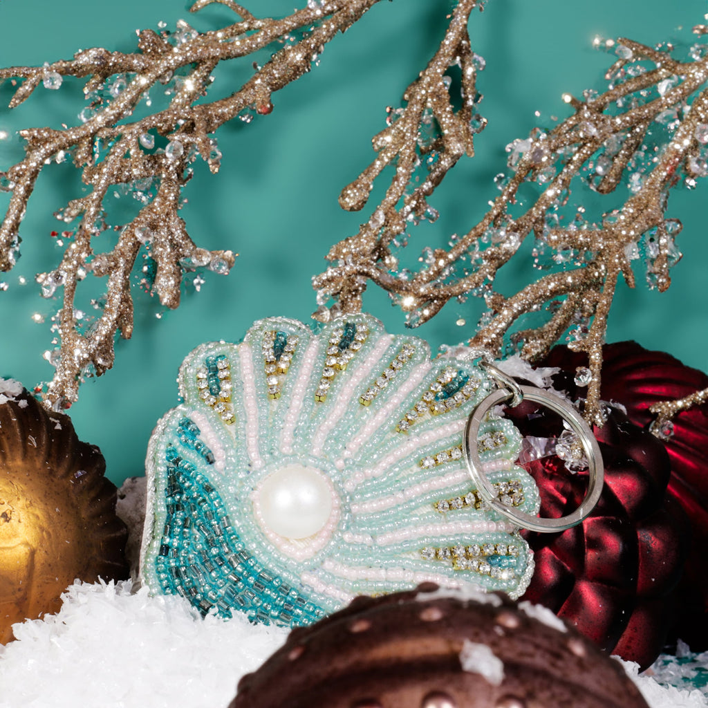 A beaded shell keyring with turquoise, pink, and gold beads and a central pearl detail resting on artificial snow among red and gold Christmas baubles and glittering gold branches against a teal background.
