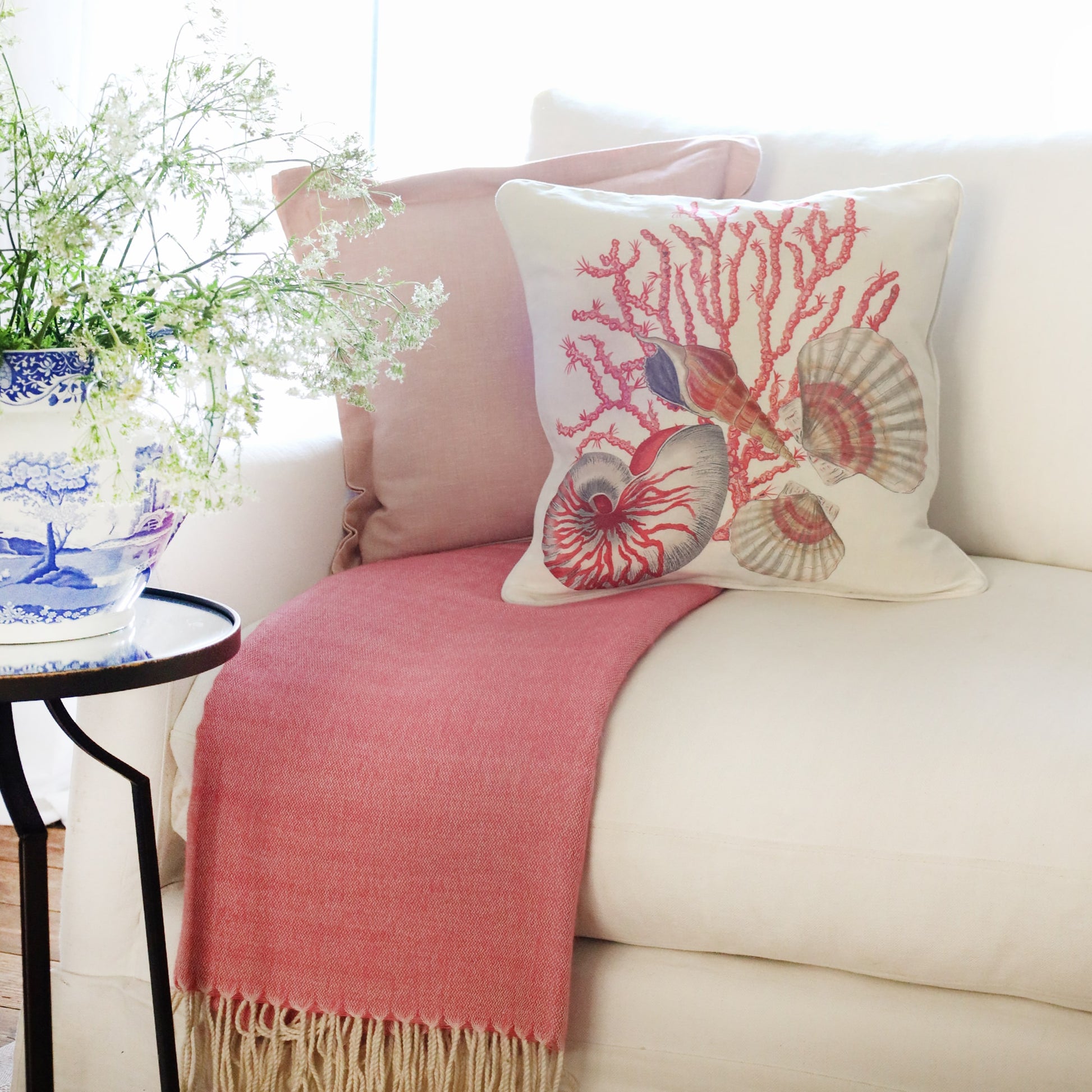 2 cushions an  a white sofa with nautilus shell, scallops and coral illustrations in tones of pink on with the sun streaming through the window at the back and a large willow pattern jug filled with cow parsley