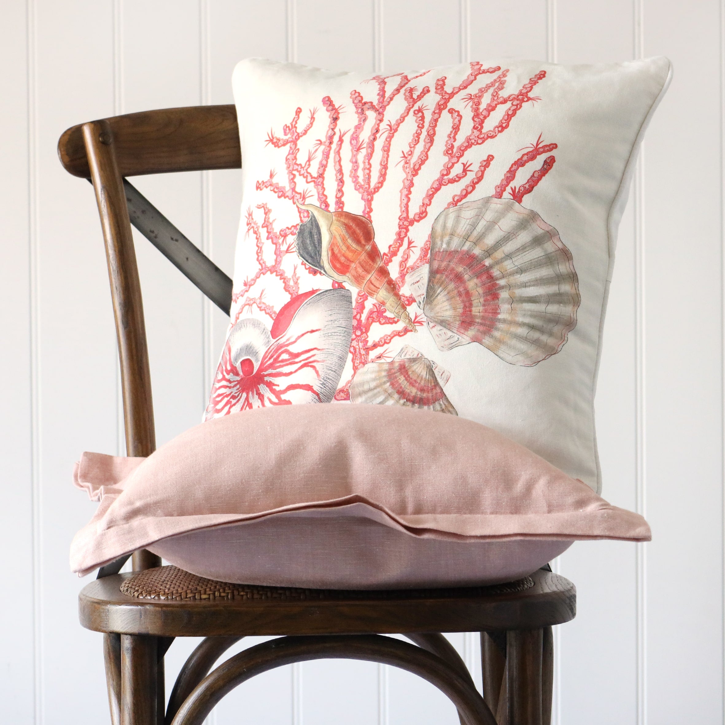 soft pink chambray cushion sitting on a wooden chair with an illustrated cushion of nautilus shell scallops and coral in tones of pink on a white ground sitting on top of it