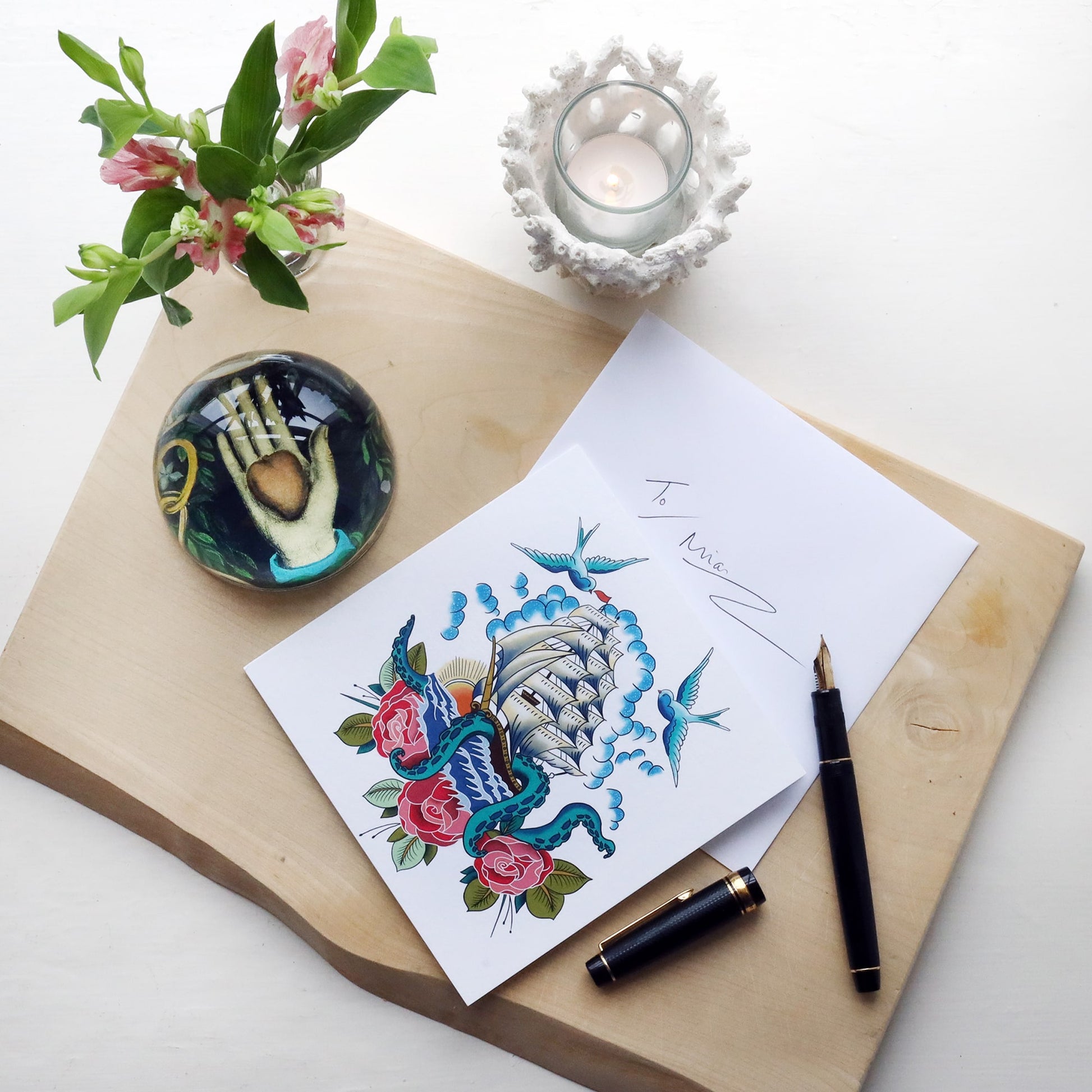 White greeting card with illustrated ship & kraken tattoo inspired design on, lying on a wooden block with fountain pen, paperweight and flowers and hand written envelope.