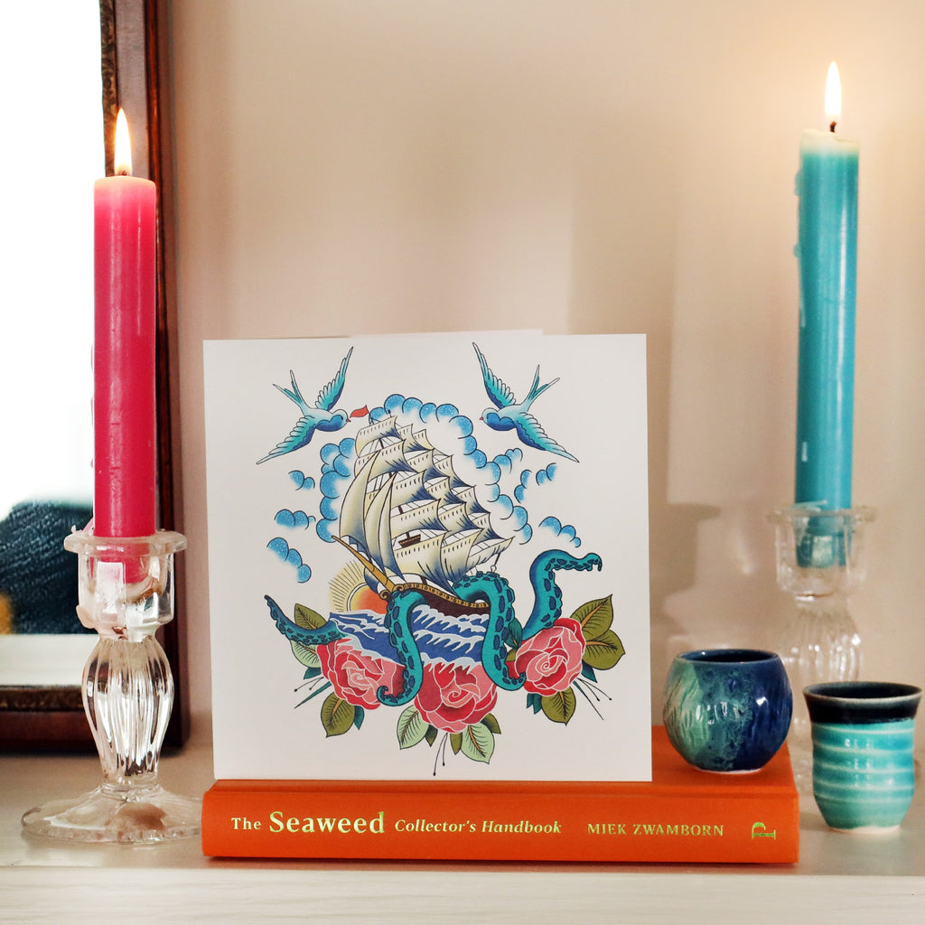 White card with illustrated ship & kraken tattoo inspired illustration, sitting on a book on a shelf with a candle lit each side and some small blue ceramic pots.