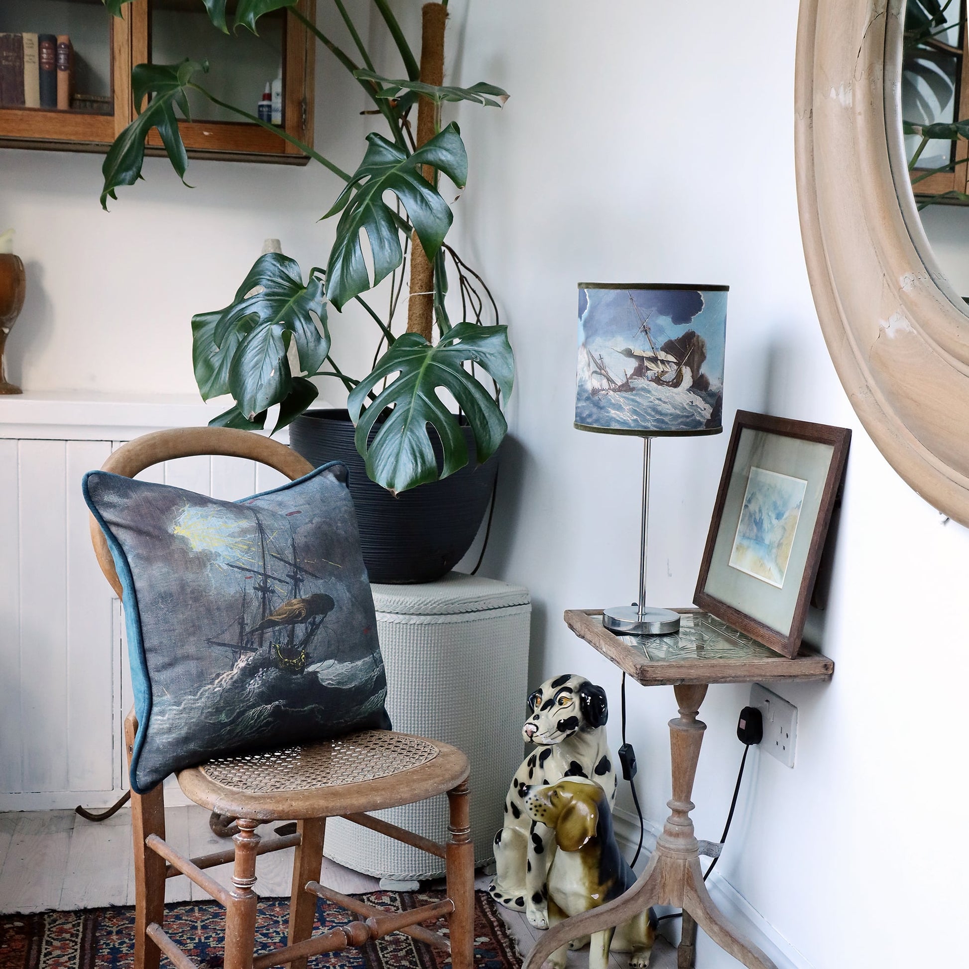 Small shipwreck scene lampshade and matching cushion set within a the corner of white room with an eclectic mix of various vintage objects and a large cheese plant.