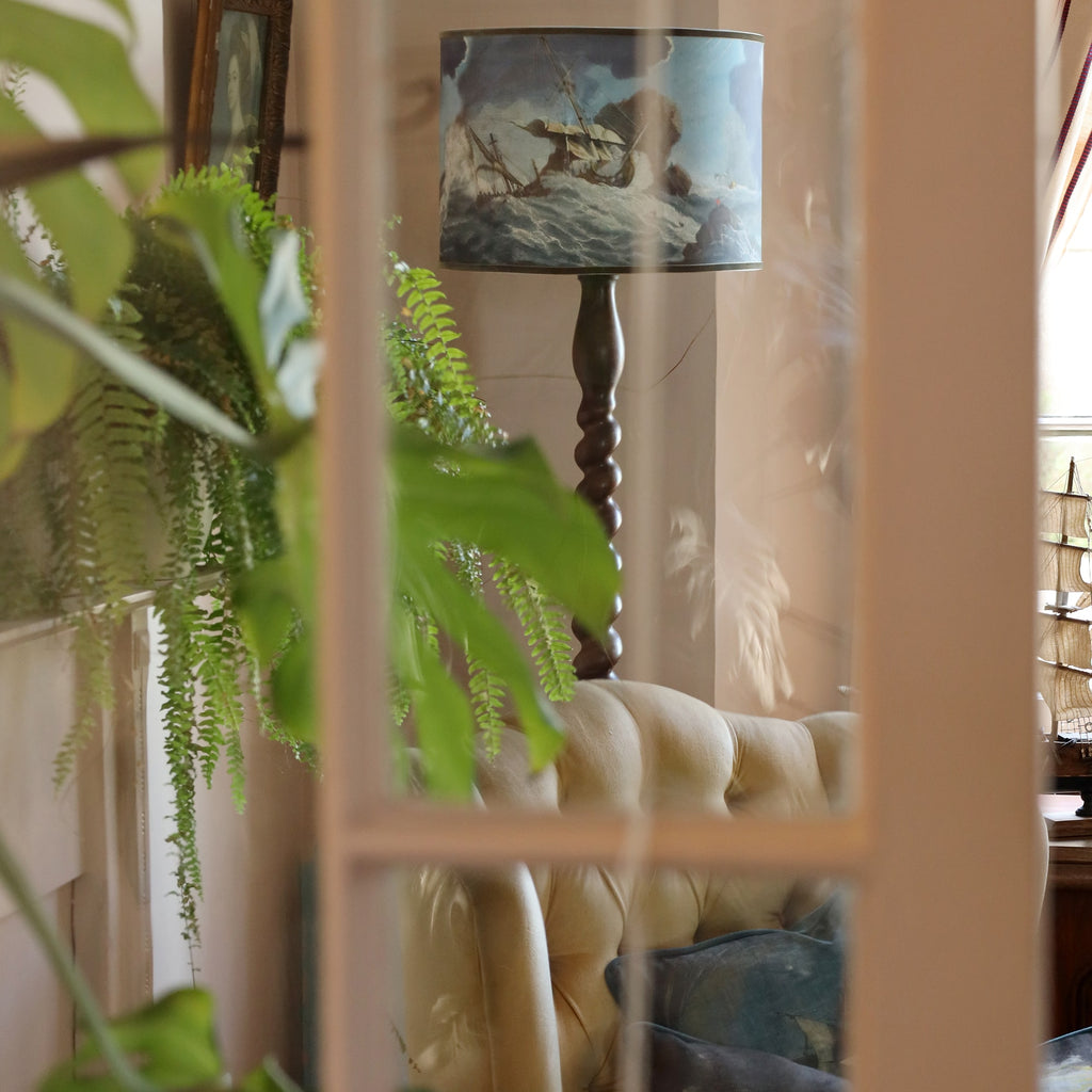 Large shipwreck scene lampshade on twisted dark wooden standard lamp base shot through a glass panelled door with refections of lots of green foliage.