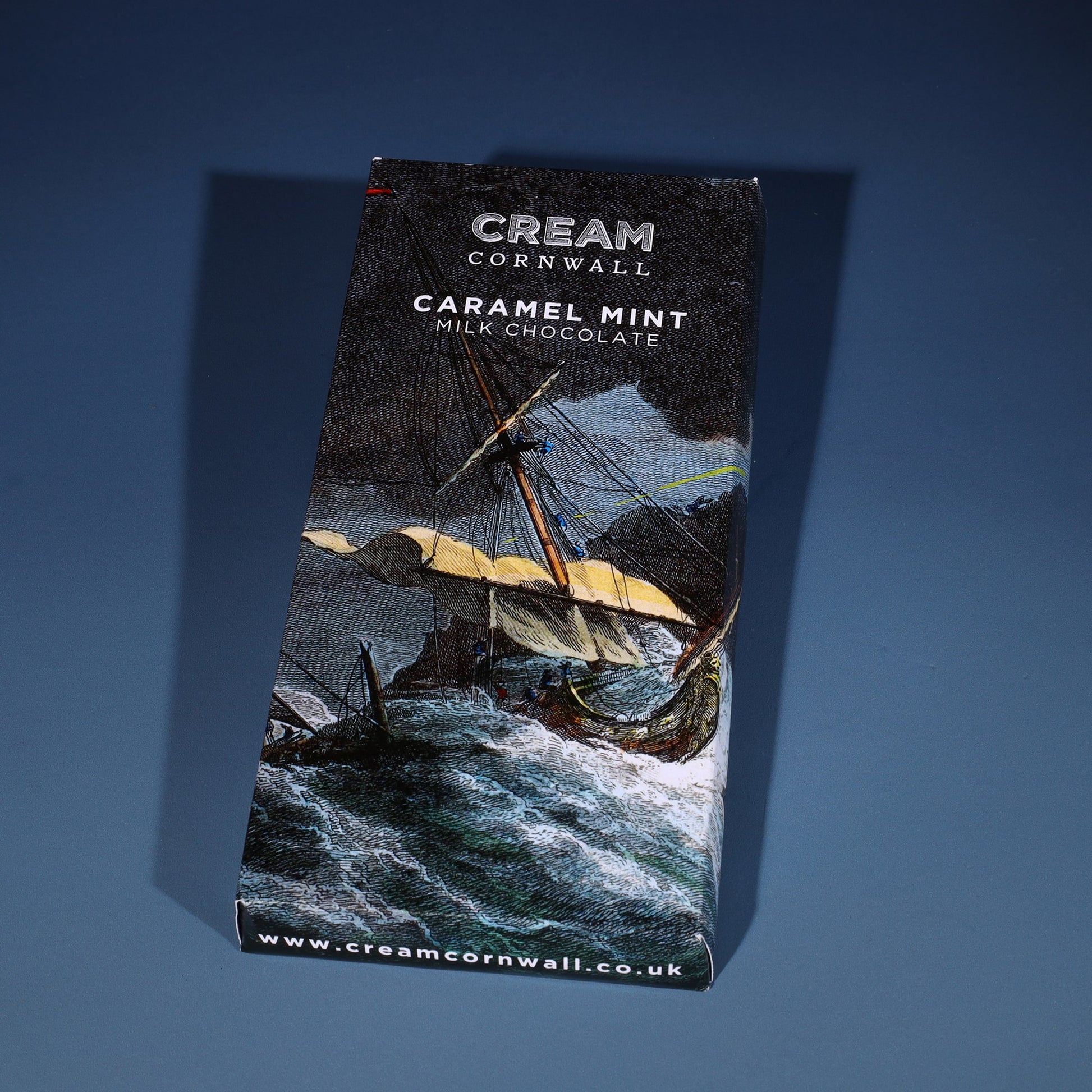 Caramel milk chocolate bar wrapped in a shipwreck scene etching. The bar has been photographed on a graduated blue background.