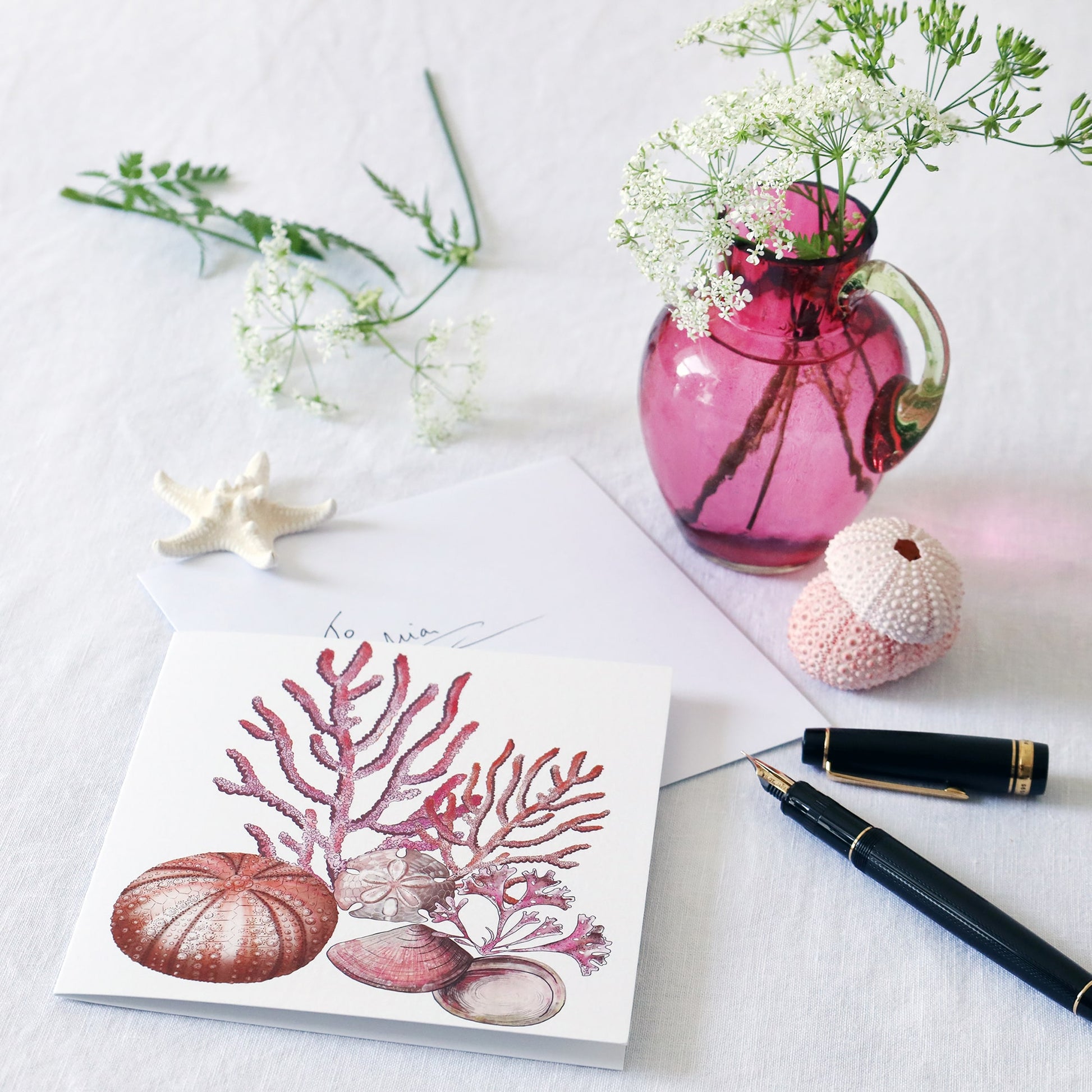 greeting card with illustration of corals, sea urchin, sand dollar and seaweed all in pinks  on a white background lying on a white table cloth with a fountain pen, hand written envelope shells and a small cranberry glass jug with wild flowers in 