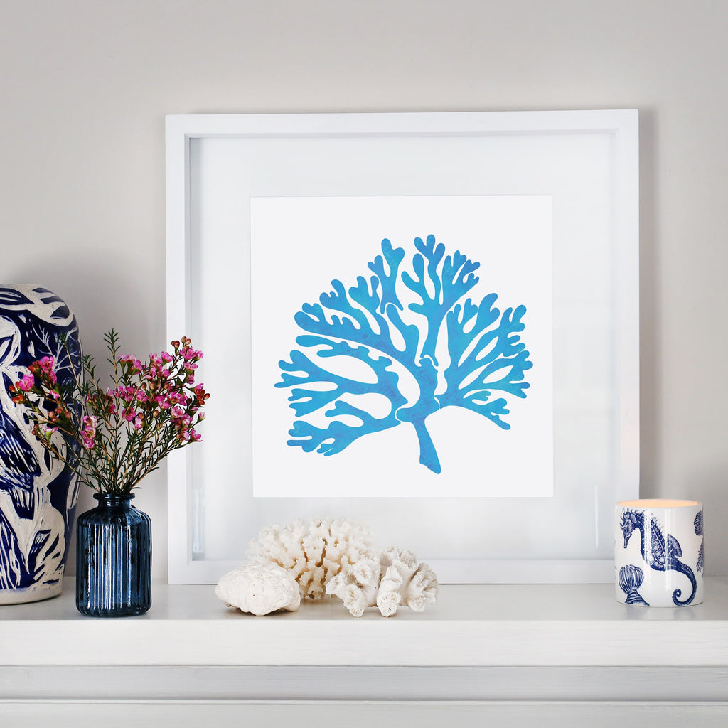A framed blue coral illustration in a white mount sits on a white mantelpiece. The display is styled with coastal-inspired decor, including a navy blue glass vase with pink flowers, white coral pieces, and a candle in a blue and white ceramic holder featuring seashell and seahorse motifs. A blue and white patterned ceramic object is partially visible on the left.