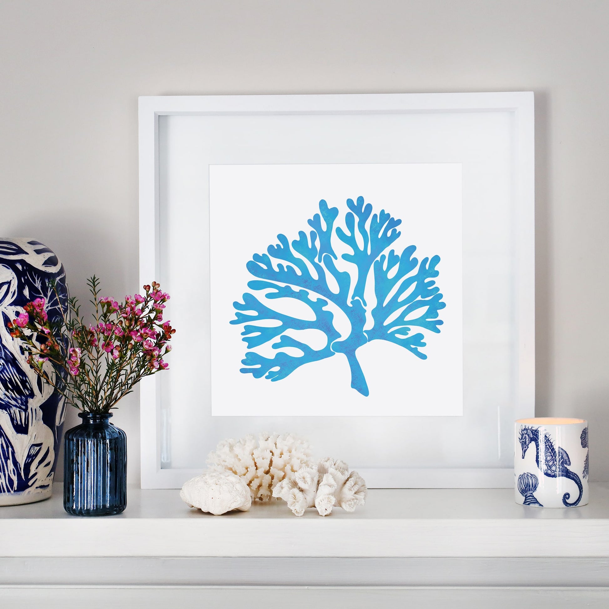 A framed blue coral illustration in a white mount sits on a white mantelpiece. The display is styled with coastal-inspired decor, including a navy blue glass vase with pink flowers, white coral pieces, and a candle in a blue and white ceramic holder featuring seashell and seahorse motifs. A blue and white patterned ceramic object is partially visible on the left.
