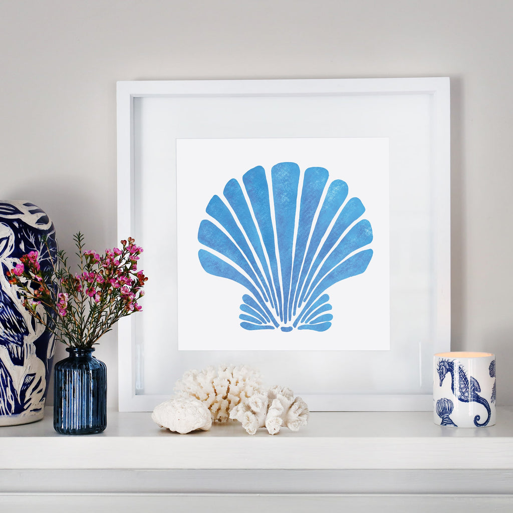 A coastal-inspired mantel display featuring a framed artwork of a bold blue scallop shell with a textured finish. The arrangement includes a small blue glass vase with pink flowers, white coral pieces, a blue and white ceramic sculpture, and a seahorse-patterned candle. The decor blends oceanic elements with soft, natural tones for a serene aesthetic.