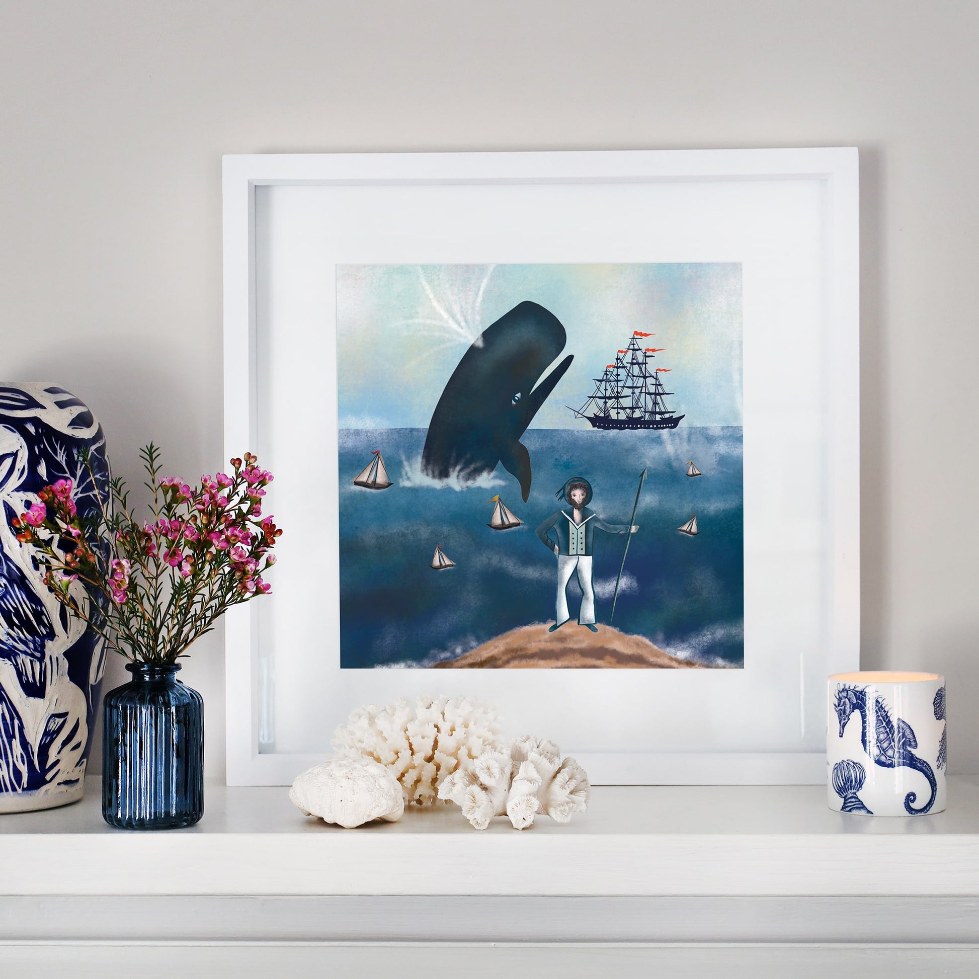 A framed nautical illustration displayed on a white mantelpiece. The artwork depicts a captain standing on a rocky shore with a spear, watching a breaching whale and a tall sailing ship surrounded by small boats. The scene is set under a misty sky. The mantel is decorated with coral, a blue glass vase with pink flowers, and a seahorse-patterned candle.