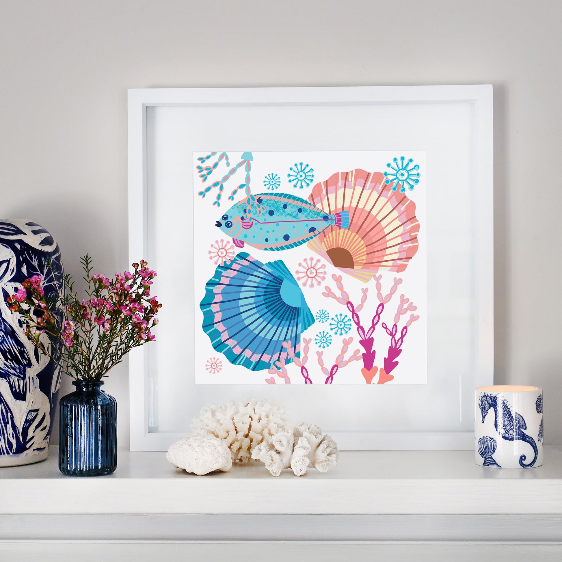 A framed illustration of a colorful flatfish swimming among vibrant seashells, coral, and sea flora in shades of blue, pink, and orange, displayed on a white mantelpiece. The setting includes decorative coastal elements such as white coral, a blue glass vase with pink flowers, a nautical-patterned ceramic cup, and a patterned sculpture, creating a harmonious ocean-inspired arrangement.