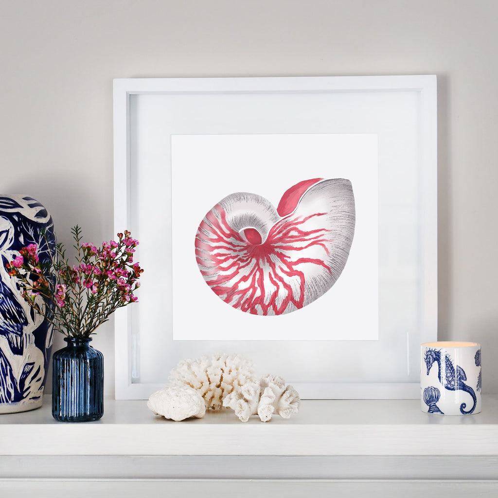 A coastal-inspired mantel display featuring a framed artwork of a detailed red and white nautilus shell. The arrangement includes a small blue glass vase with pink flowers, white coral pieces, a blue and white ceramic sculpture, and a seahorse-patterned candle. The decor blends oceanic elements with soft, natural tones for a serene aesthetic.