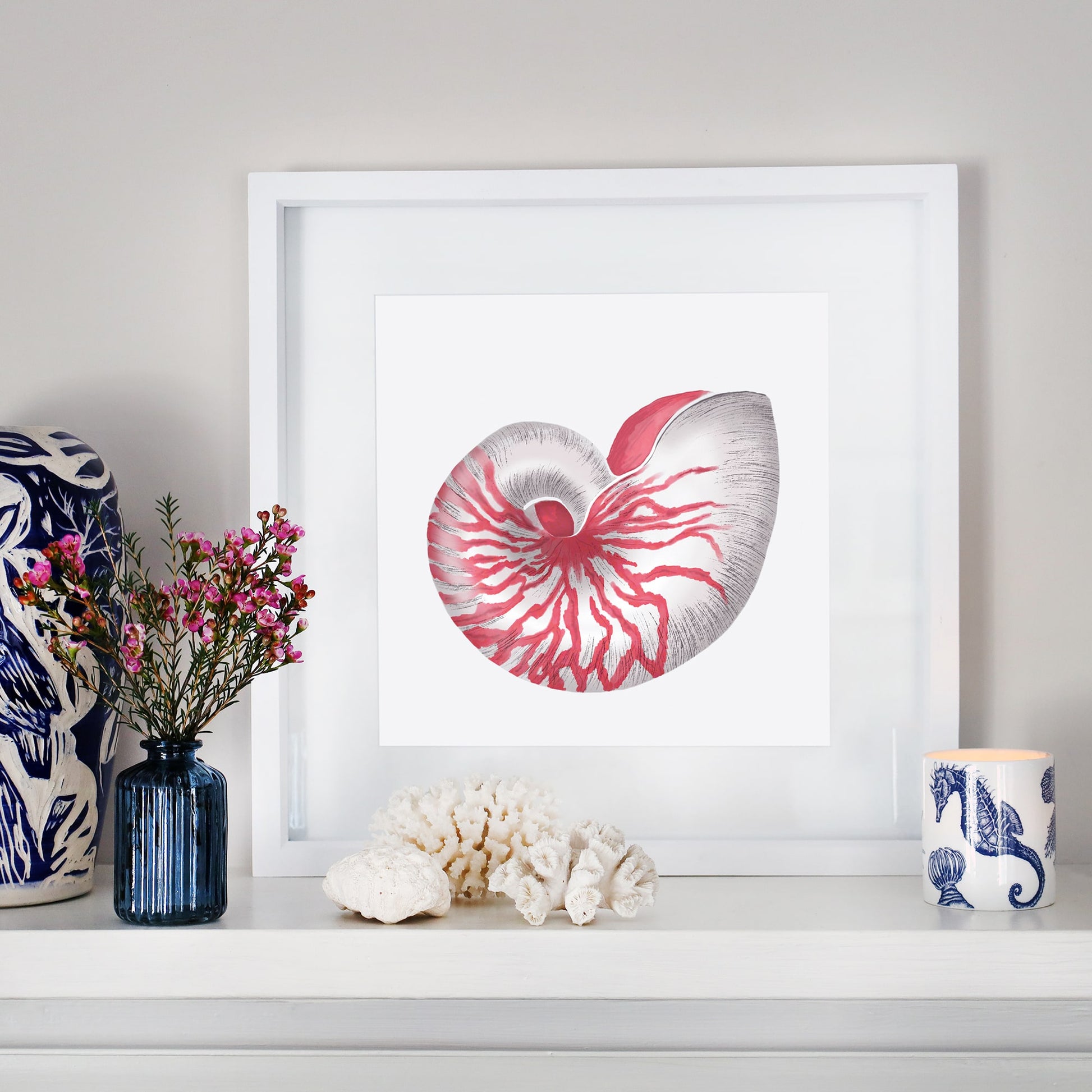 A coastal-inspired mantel display featuring a framed artwork of a detailed red and white nautilus shell. The arrangement includes a small blue glass vase with pink flowers, white coral pieces, a blue and white ceramic sculpture, and a seahorse-patterned candle. The decor blends oceanic elements with soft, natural tones for a serene aesthetic.