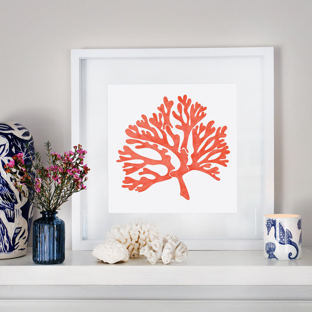 A coastal-inspired mantelpiece featuring a framed orange coral illustration in a white frame. The display includes decorative elements such as a blue vase with pink flowers, white coral pieces, and a blue and white candle holder with a seahorse design, creating a serene, beach-themed aesthetic.