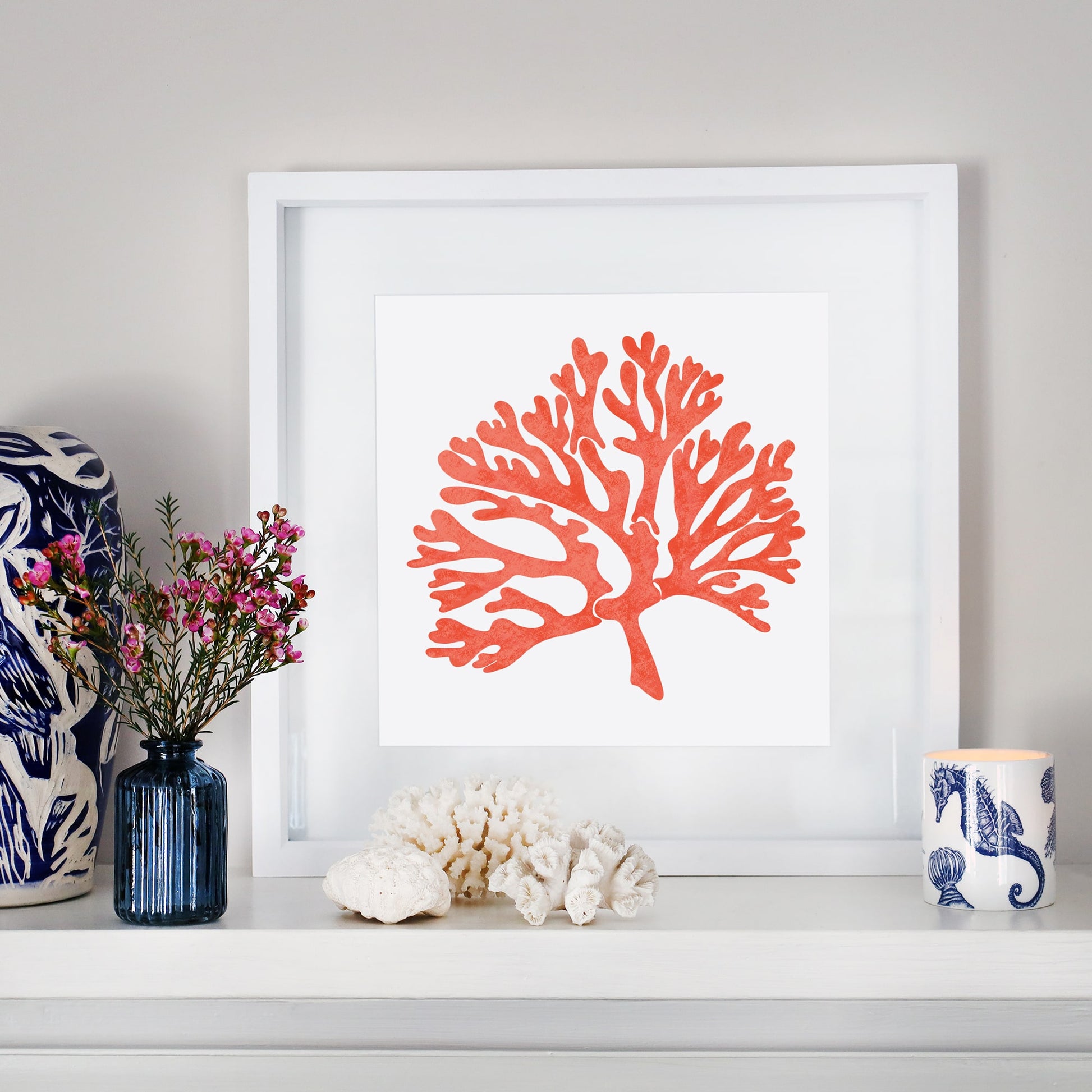 A coastal-inspired mantelpiece featuring a framed orange coral illustration in a white frame. The display includes decorative elements such as a blue vase with pink flowers, white coral pieces, and a blue and white candle holder with a seahorse design, creating a serene, beach-themed aesthetic.