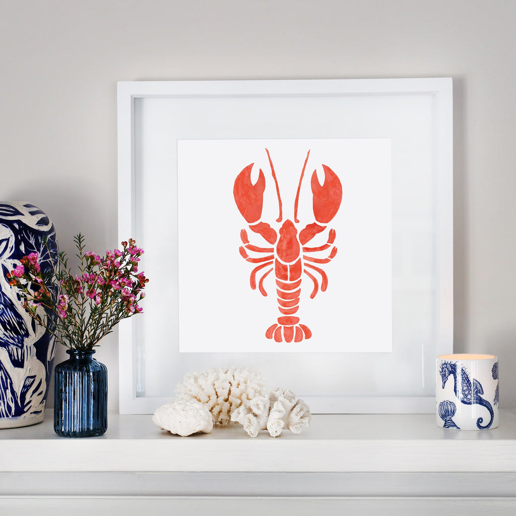 A framed illustration of an orange lobster is displayed on a white mantelpiece, surrounded by coastal-themed decor, including coral, seashells, a blue vase with pink flowers, and a candle in a blue and white nautical-patterned holder.