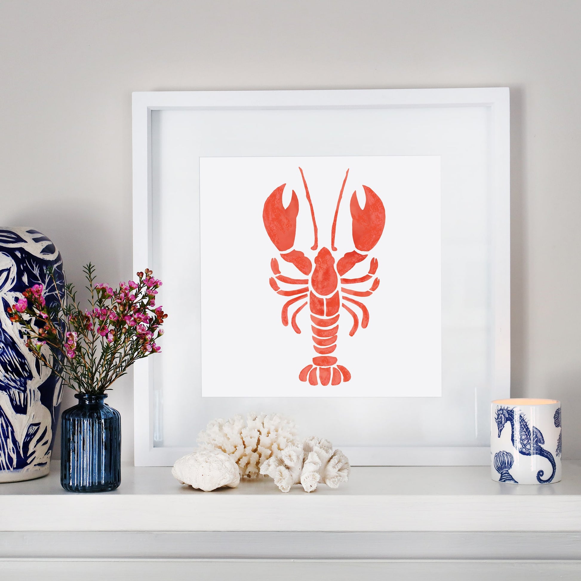 A framed illustration of an orange lobster is displayed on a white mantelpiece, surrounded by coastal-themed decor, including coral, seashells, a blue vase with pink flowers, and a candle in a blue and white nautical-patterned holder.