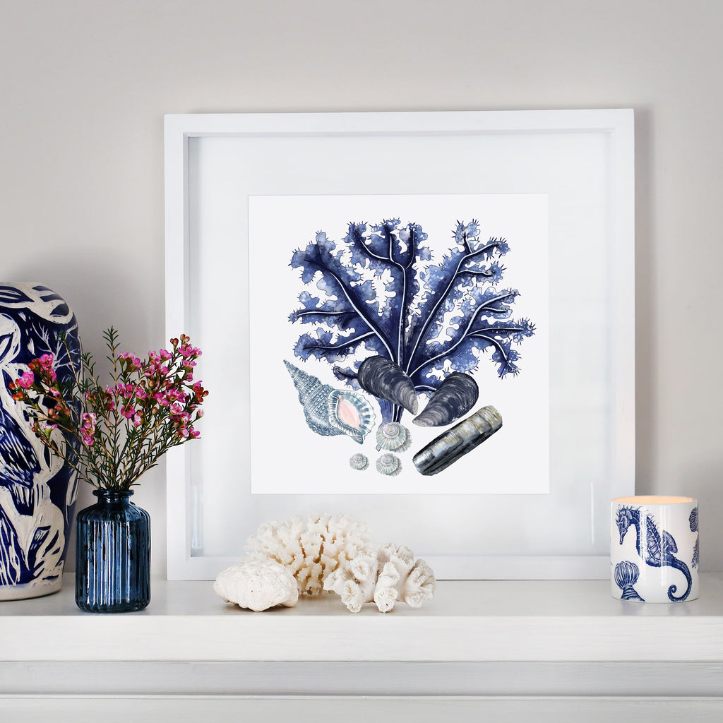 Framed coastal print of blue coral and seashells on a white mantelpiece, styled with coral pieces, a blue glass vase with flowers, and a nautical-themed candle holder.