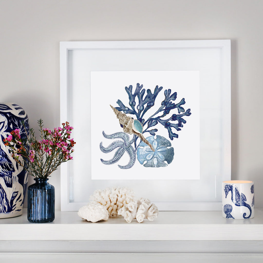 A coastal-inspired mantel display featuring a framed artwork of blue coral, a seashell, a starfish, and a sand dollar. The arrangement includes a small blue glass vase with pink flowers, white coral pieces, a blue and white ceramic sculpture, and a seahorse-patterned candle. The decor blends oceanic elements with soft, natural tones for a serene aesthetic.