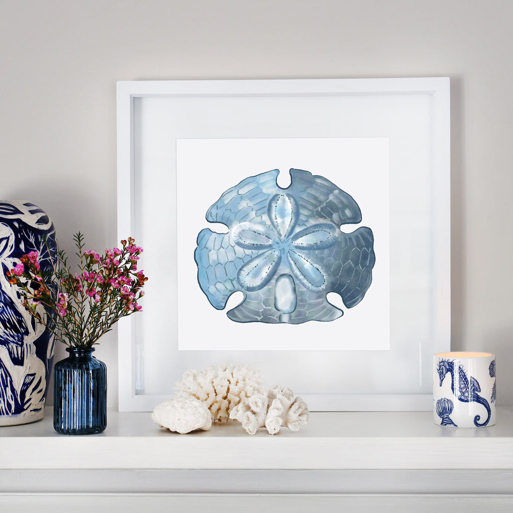 A coastal-inspired mantel display featuring a framed artwork of a detailed blue sand dollar. The arrangement includes a small blue glass vase with pink flowers, white coral pieces, a blue and white ceramic sculpture, and a seahorse-patterned candle. The decor blends oceanic elements with soft, natural tones for a serene aesthetic.