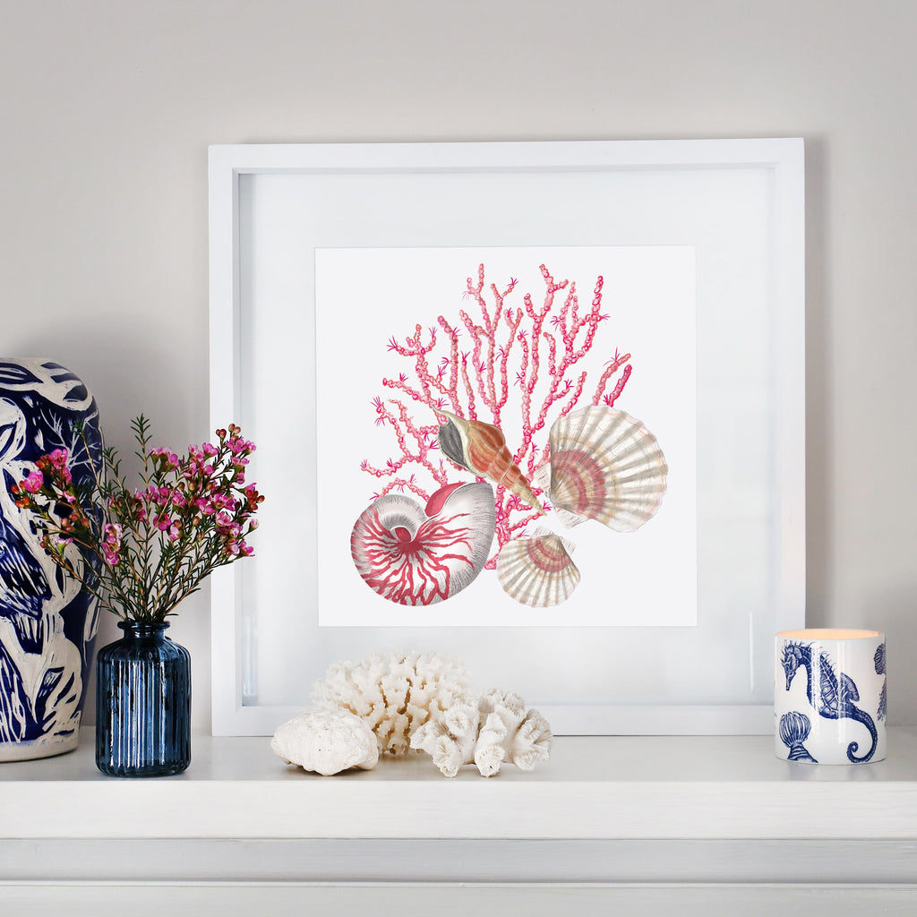A coastal-inspired mantel display featuring a framed artwork of pink coral, a nautilus shell, scallop shells, and a spiral seashell. The arrangement includes a small blue glass vase with pink flowers, white coral pieces, a blue and white ceramic sculpture, and a seahorse-patterned candle. The decor blends oceanic elements with soft, natural tones for a serene aesthetic.